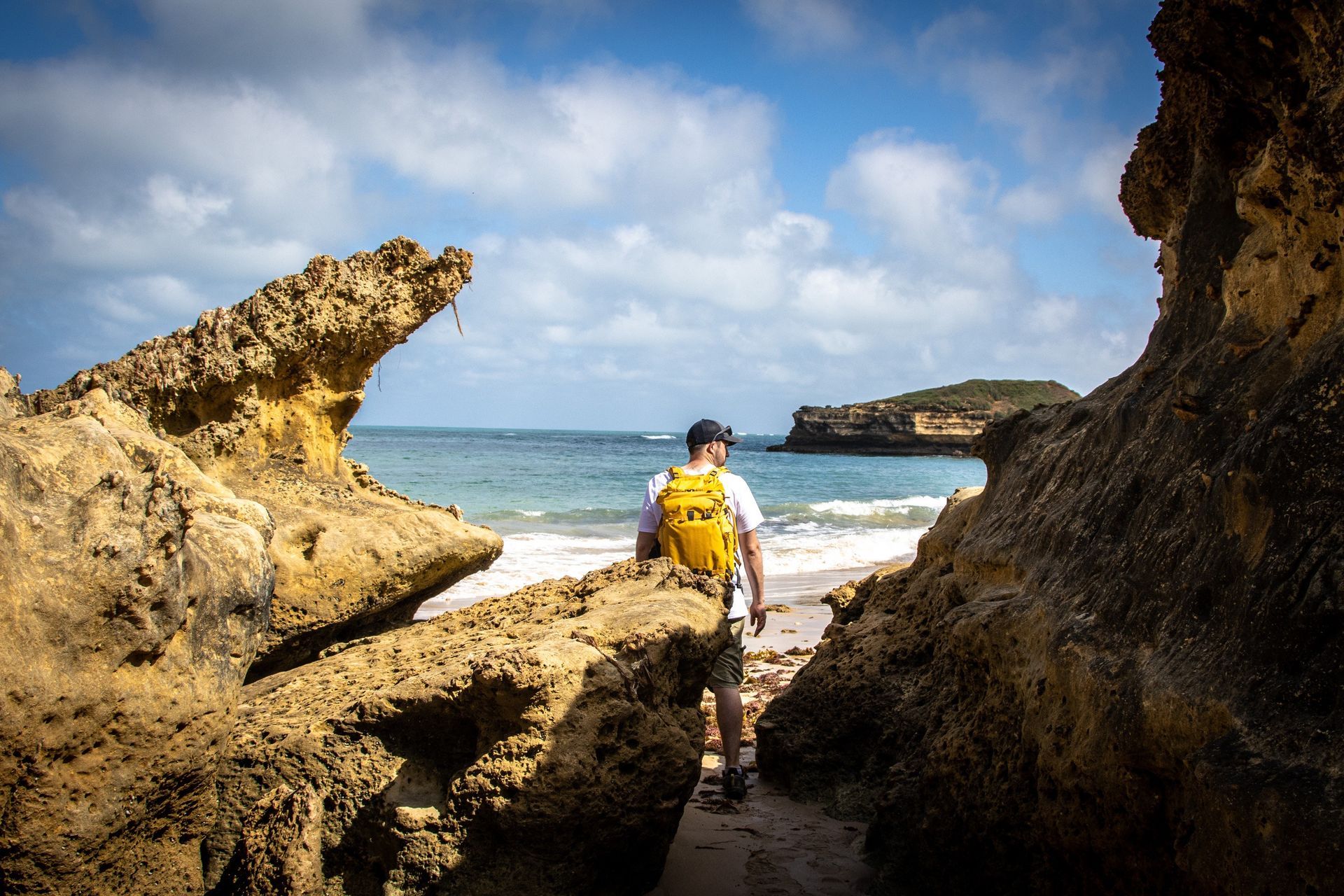 Man with yellow backpack walks towards beach, framed by rock formations. Blue sky, ocean.