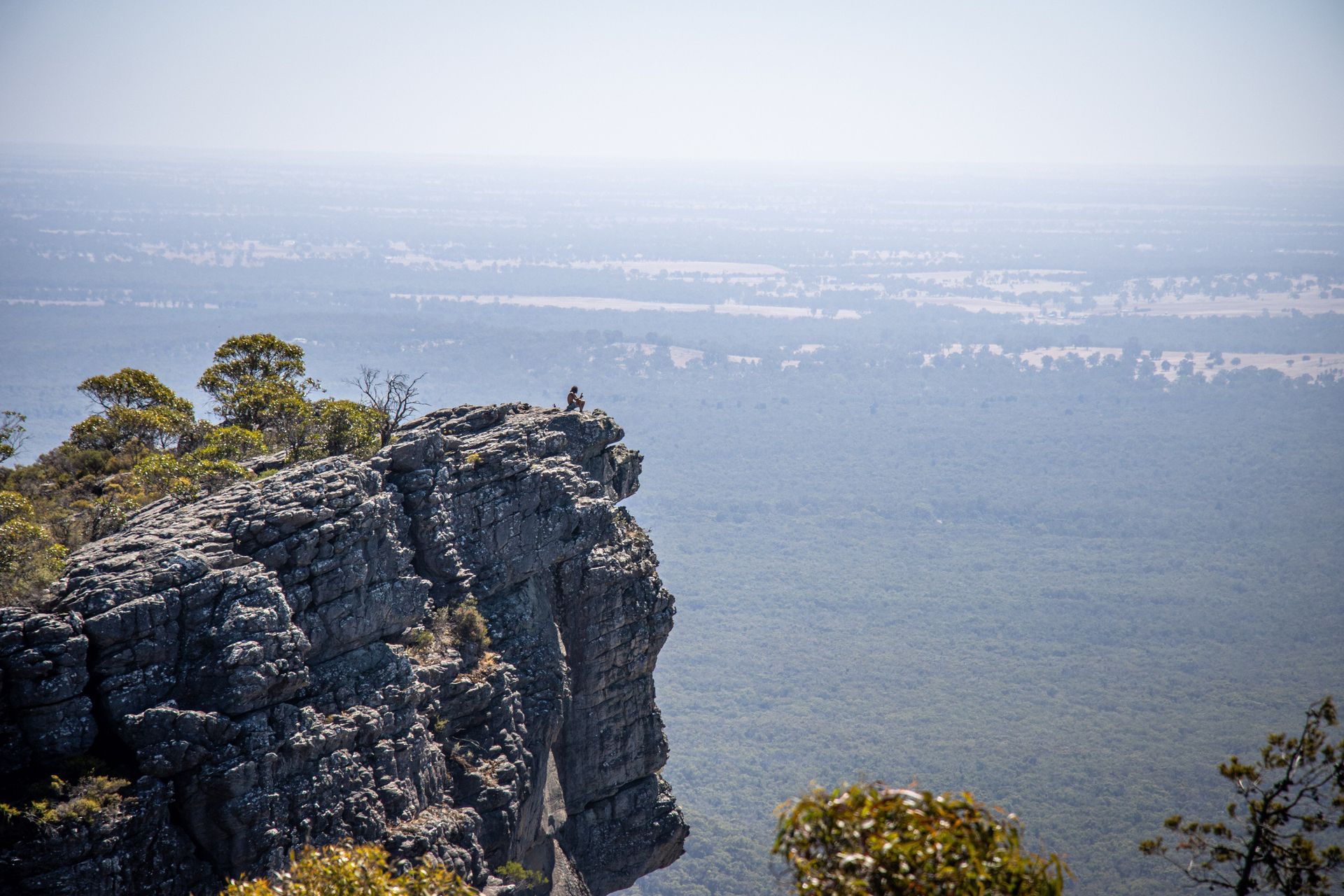 Person sitting on a cliff edge overlooking a vast forested valley on a hazy day.