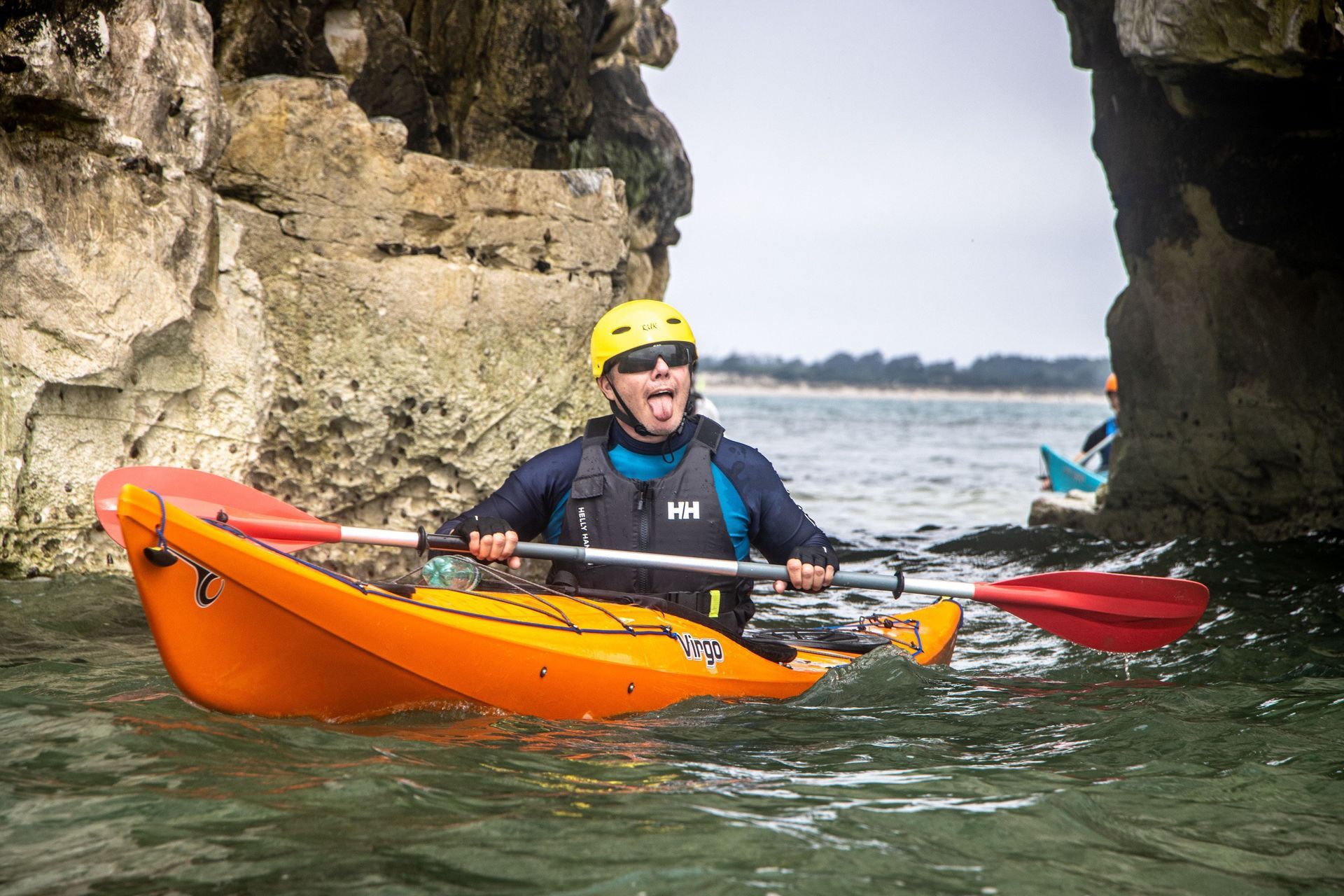 Man in orange kayak sticking out tongue, passing through a rock archway, holding paddle.