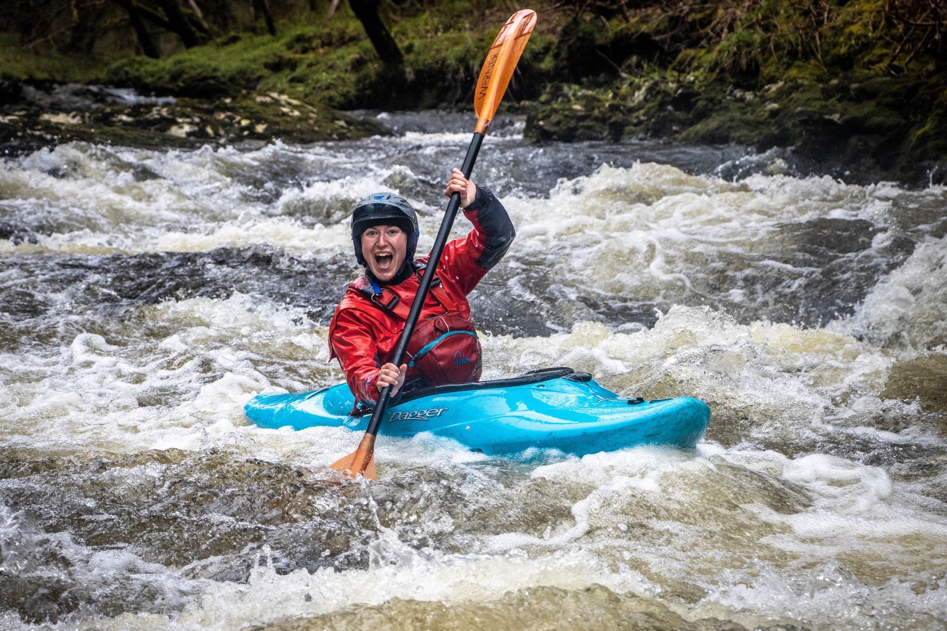 Kayaker in red gear smiles, paddles in rushing whitewater, surrounded by brown river.