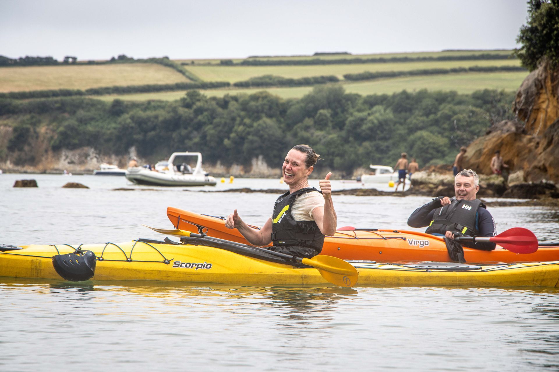 Two people in kayaks on water, one giving thumbs up. Boats and shoreline in the background.
