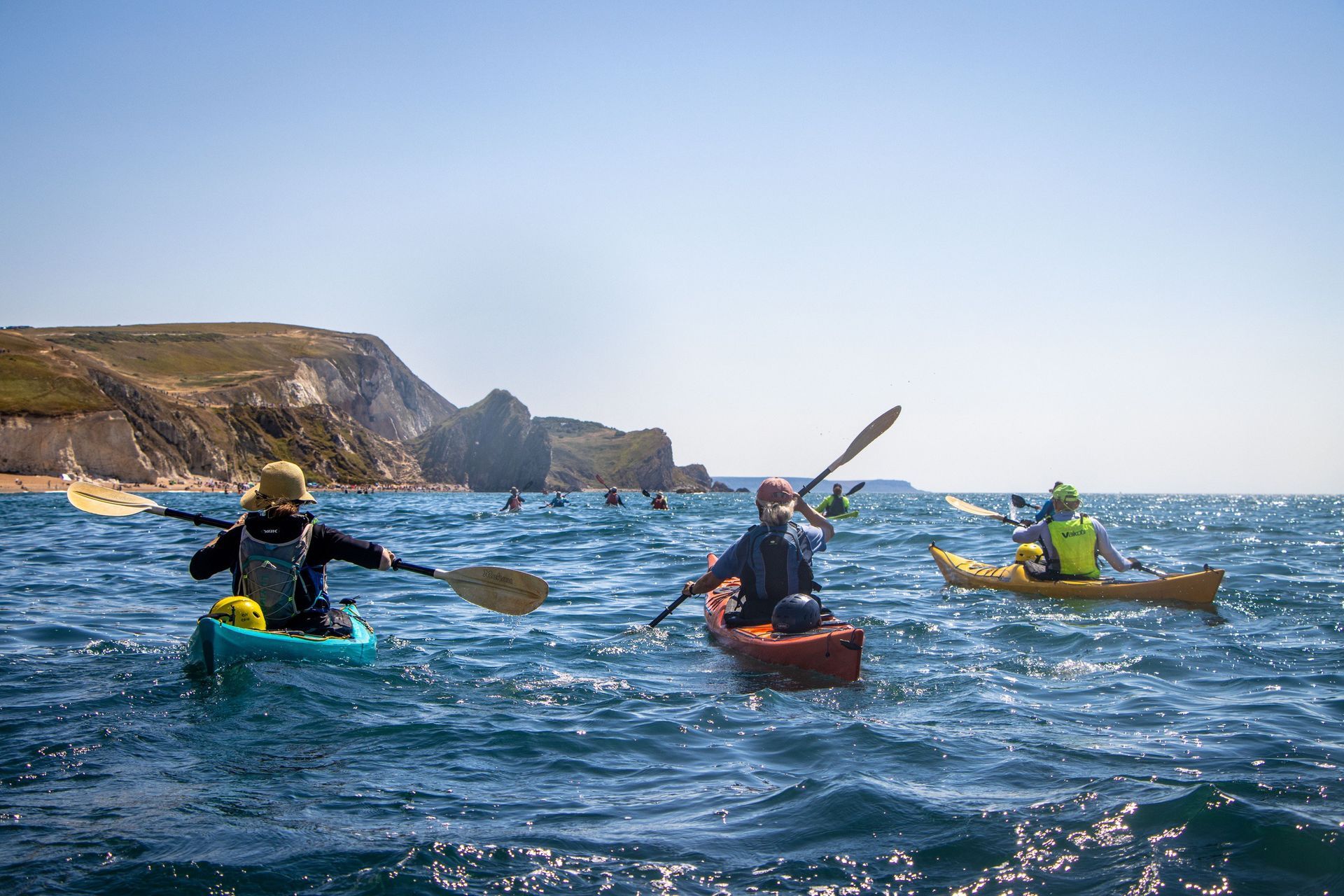Kayakers paddle on blue water near white cliffs under a clear sky.