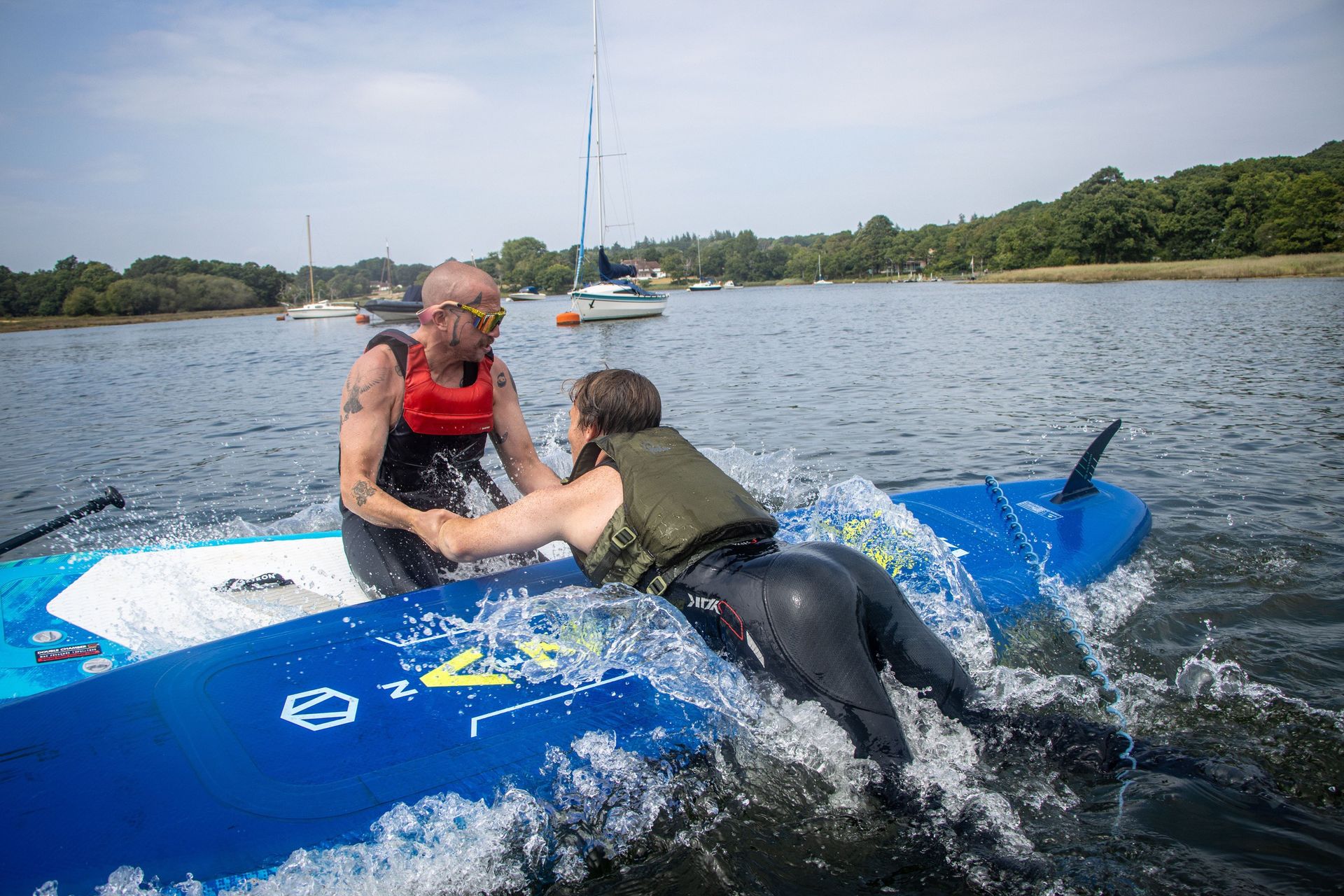 Man helping another onto a blue paddleboard in water, both wearing life vests. Boats in background.