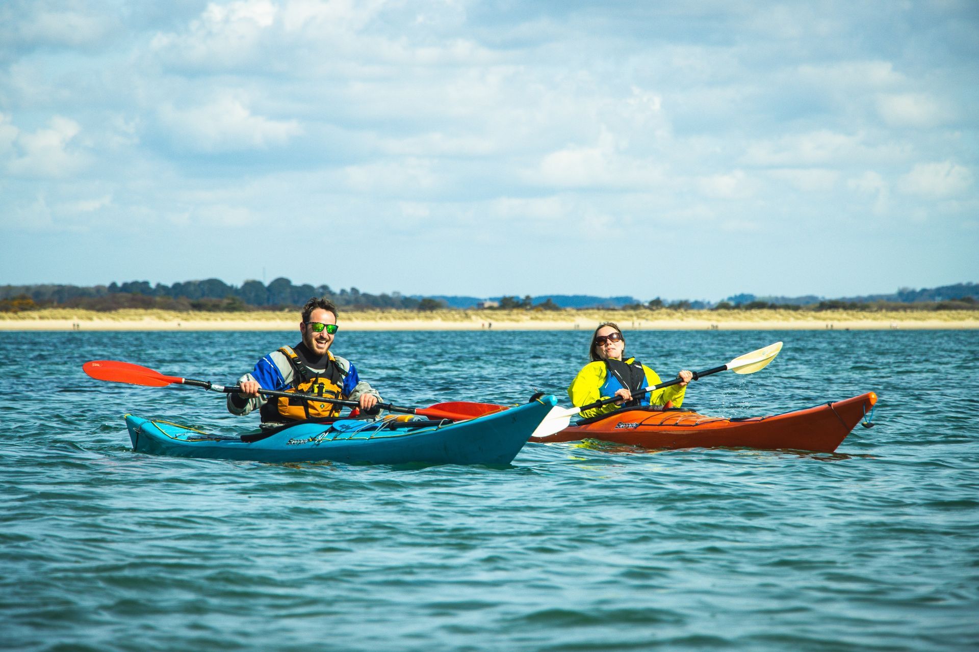 Two people kayaking in calm blue water. One wears a blue life vest, the other yellow. Sunny day.