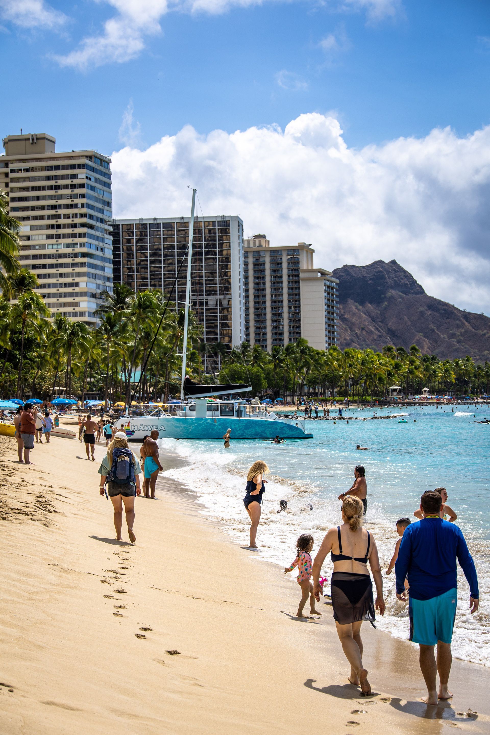 Beach scene with people walking, swimming, and enjoying the sun. Buildings and Diamond Head visible in the background.
