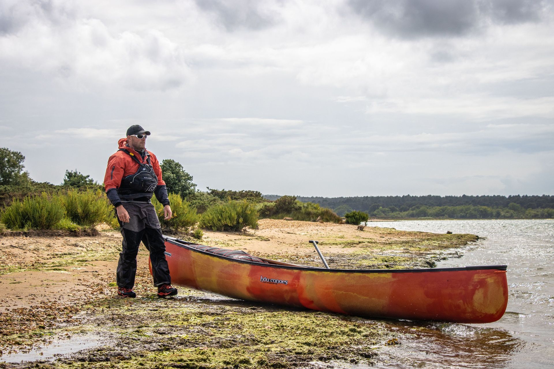 Man in rain gear stands near canoe on a shoreline, overcast sky in background.