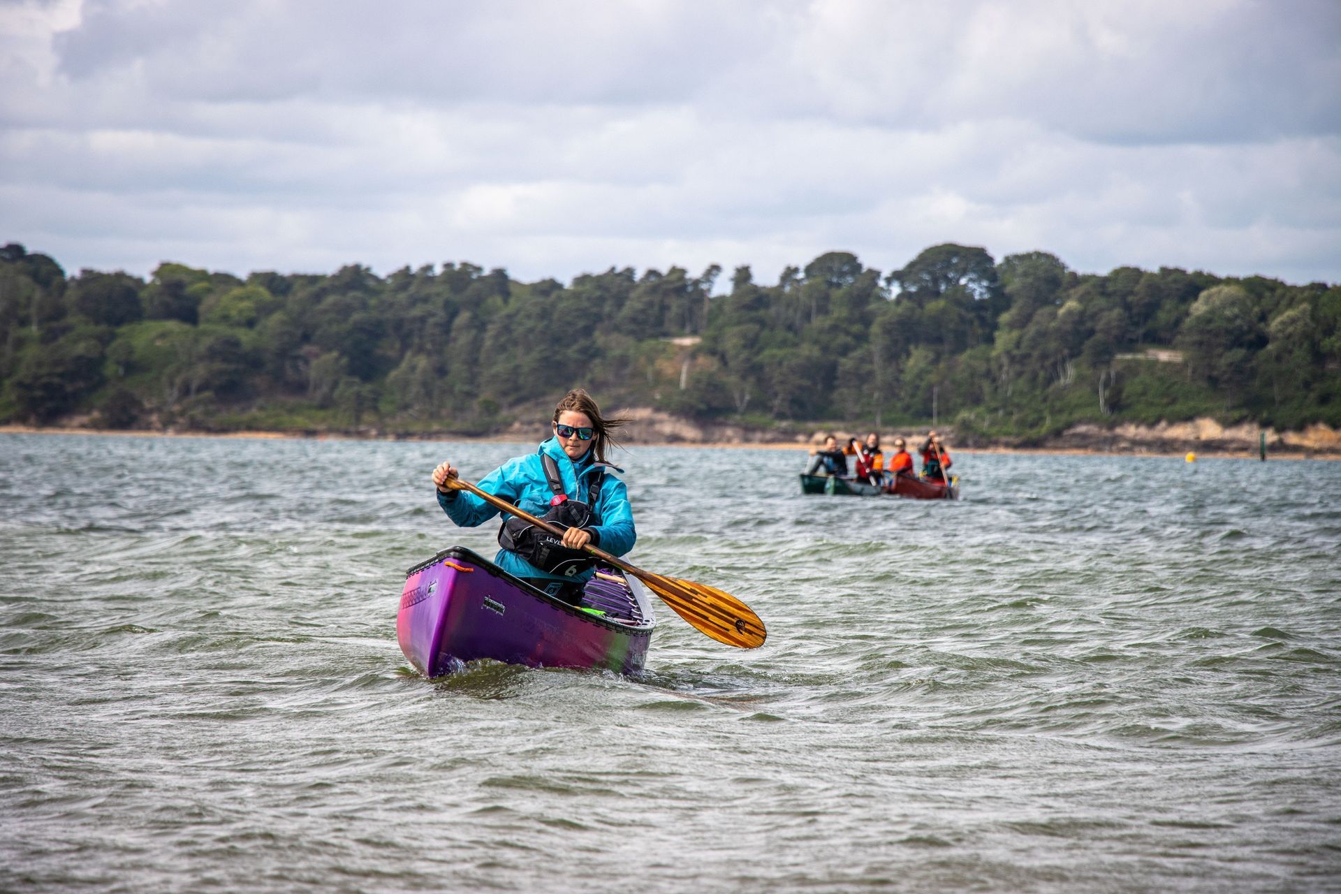 Person canoeing in a purple boat on water, others in a canoe behind, trees in background. Cloudy sky.