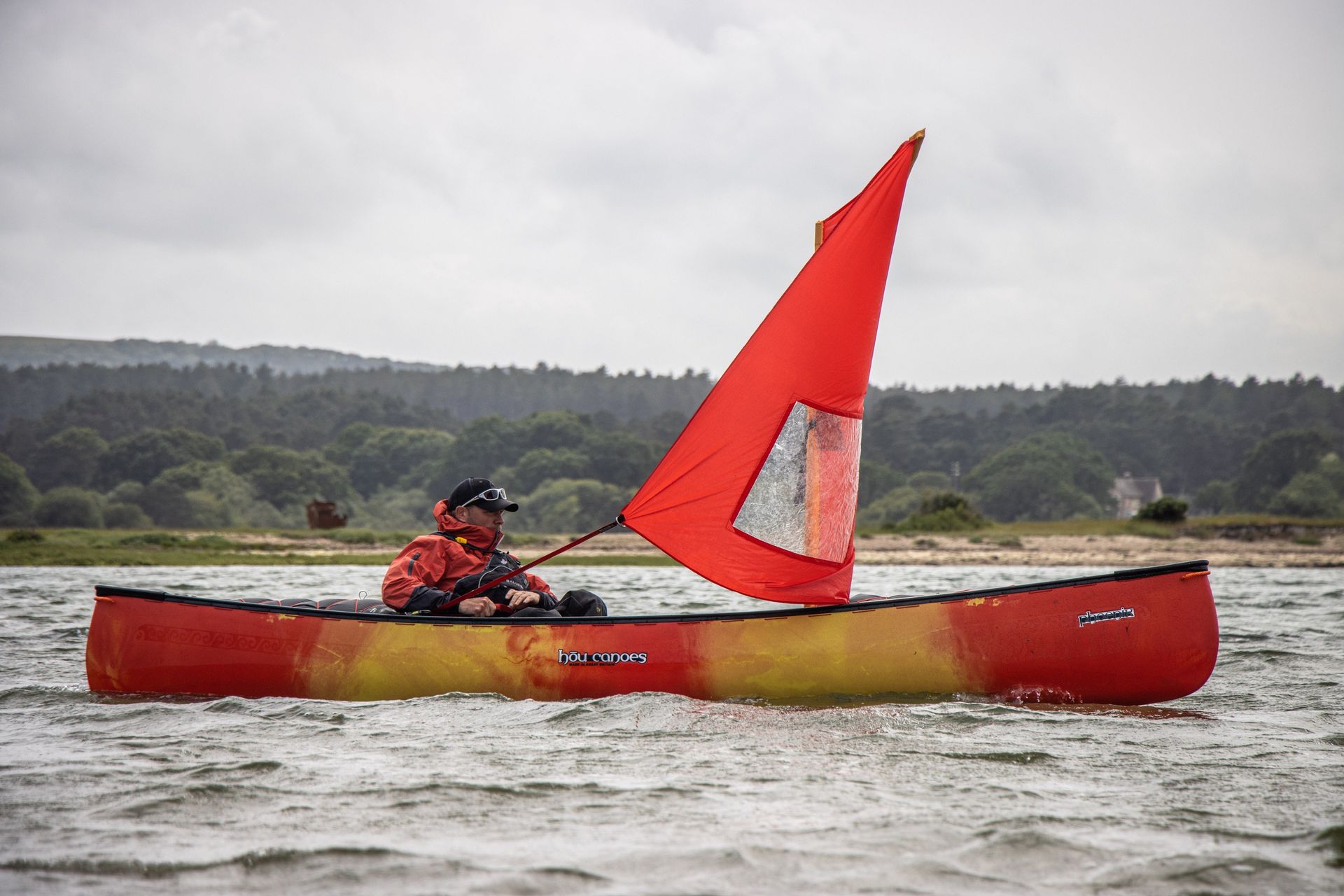 Person sails a red canoe with a red sail on a body of water, cloudy day.