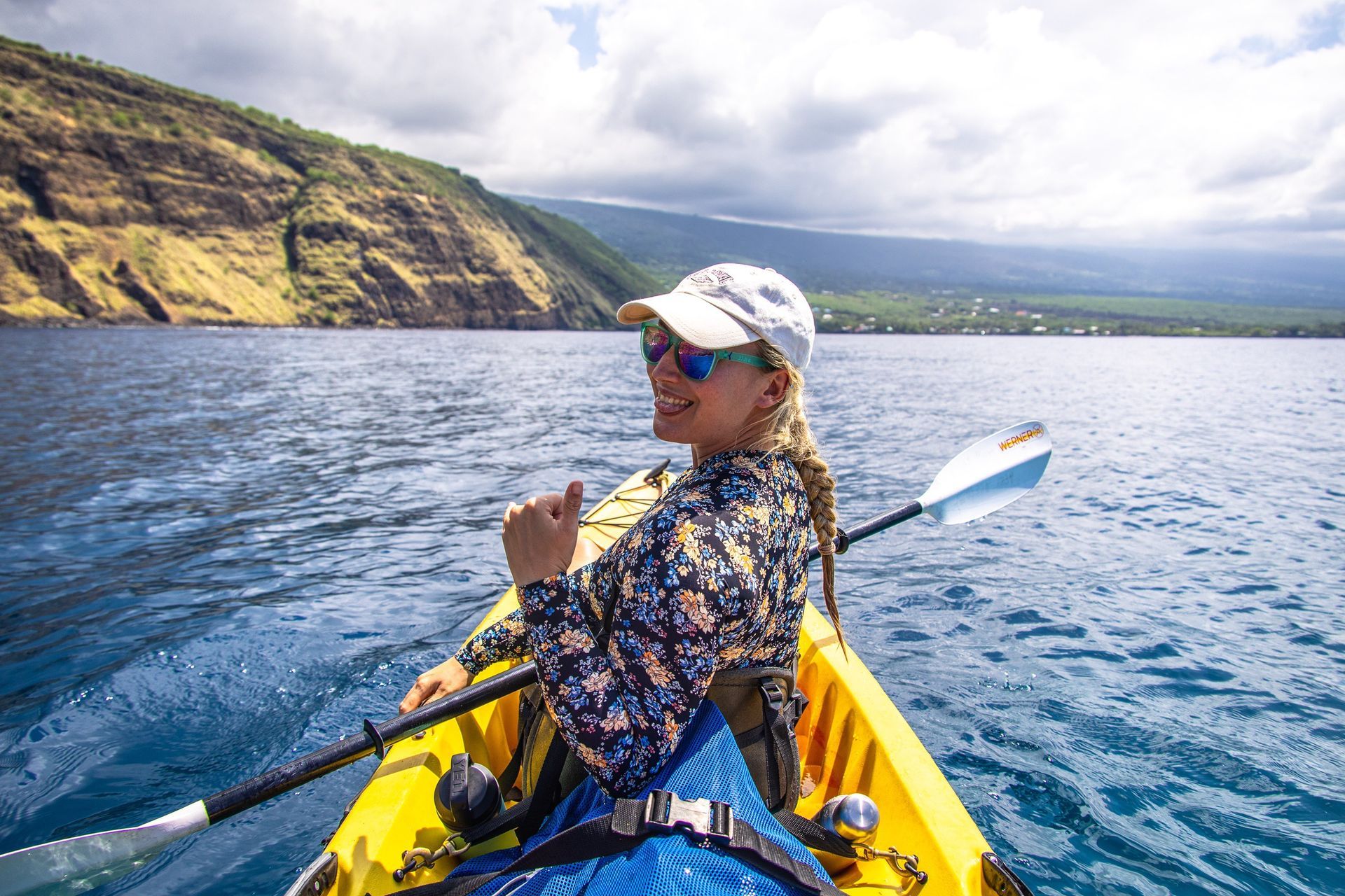 Woman kayaking in a yellow kayak, smiling, thumbs up, ocean, mountains, cloudy sky.