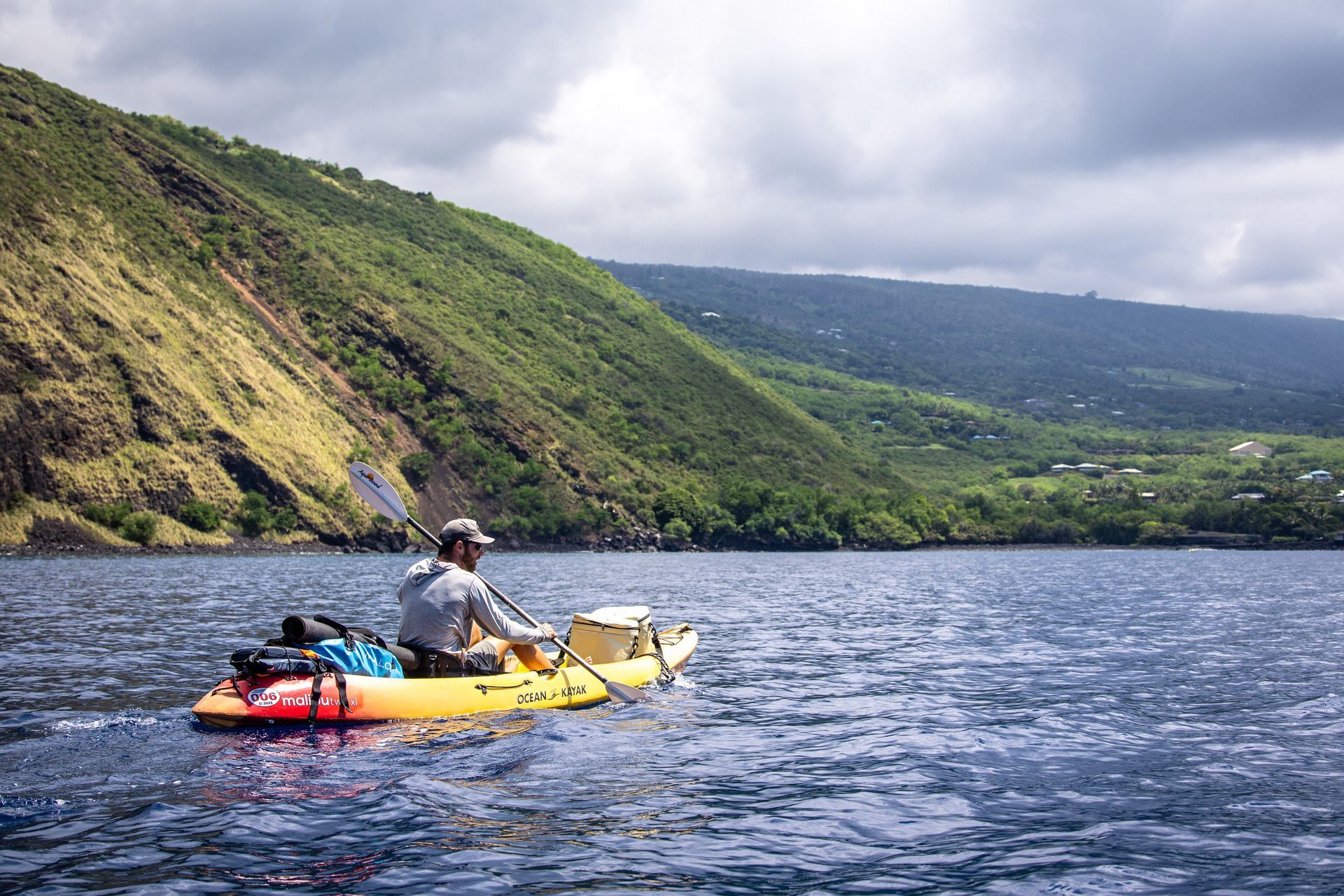 Man paddles a yellow kayak on blue water near a lush green hillside.