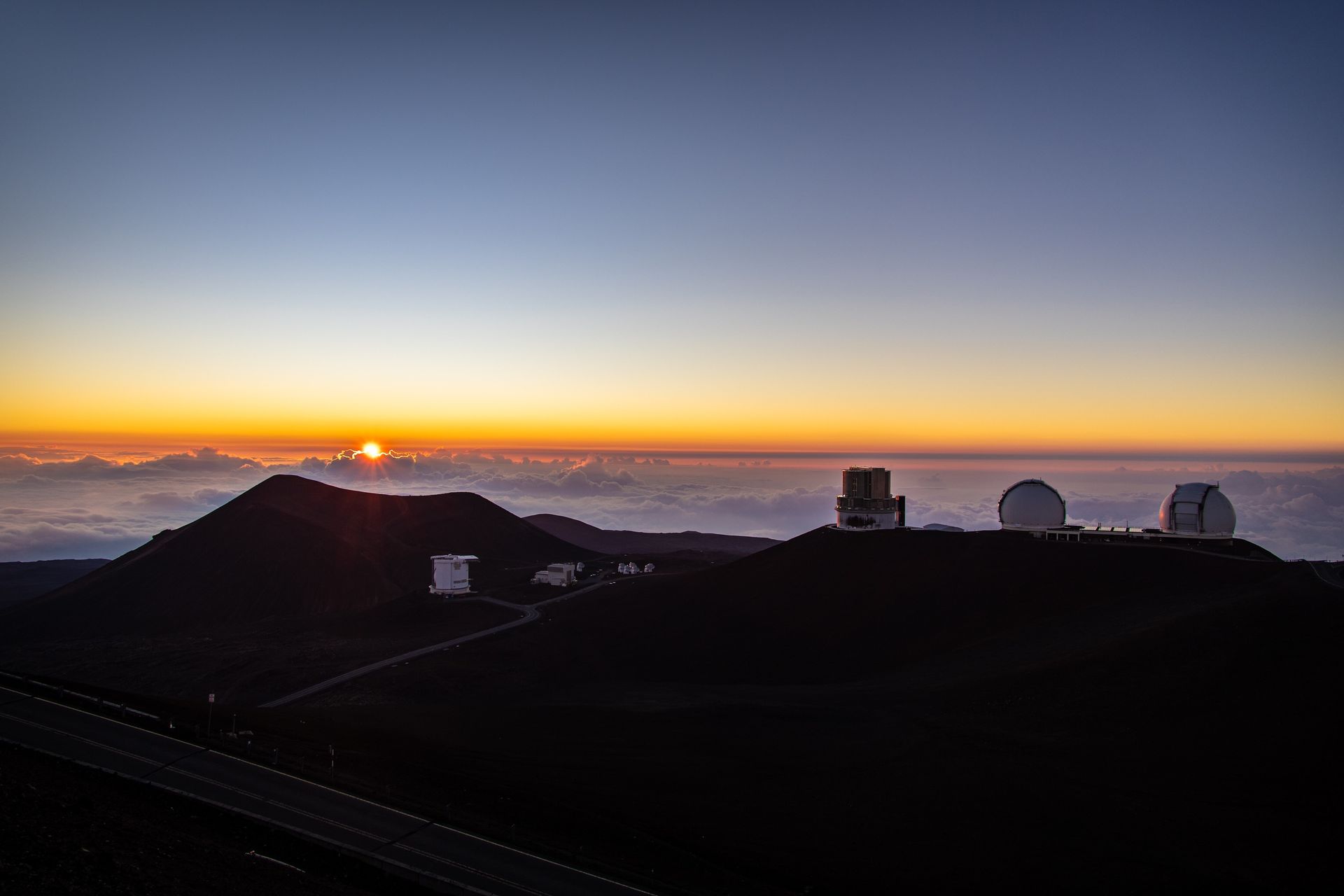 Observatory domes atop a mountain at sunset, with a road in the foreground and golden light illuminating the sky.