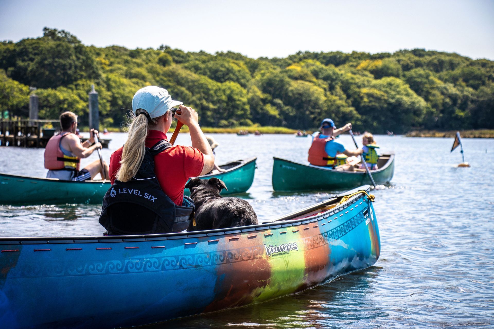 People canoeing on a sunny lake. A woman in a red shirt with a dog, others paddling, and a green shoreline.