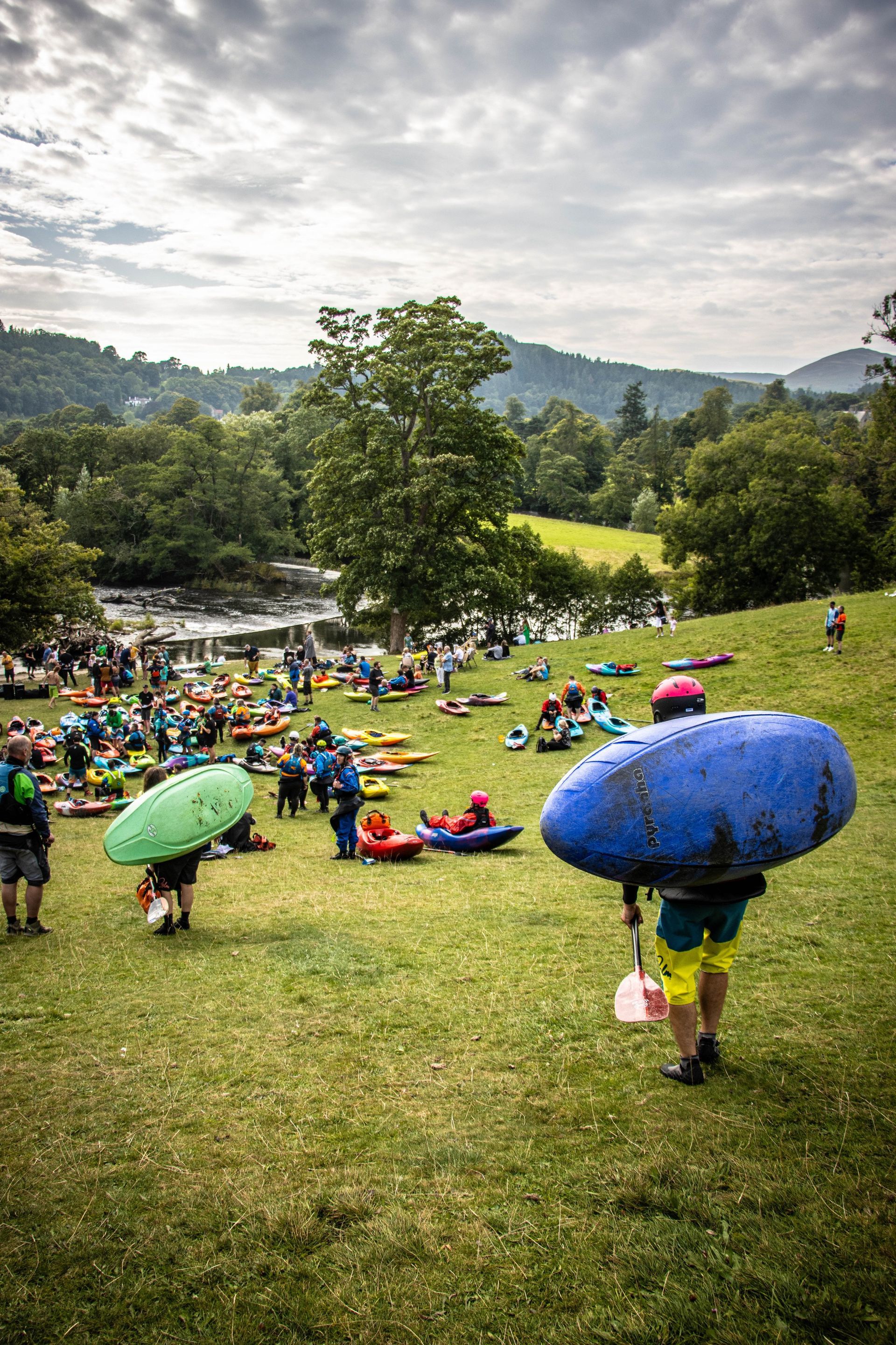 People carrying inflatable rafts on a grassy bank near a river, with hills and overcast sky in the background.