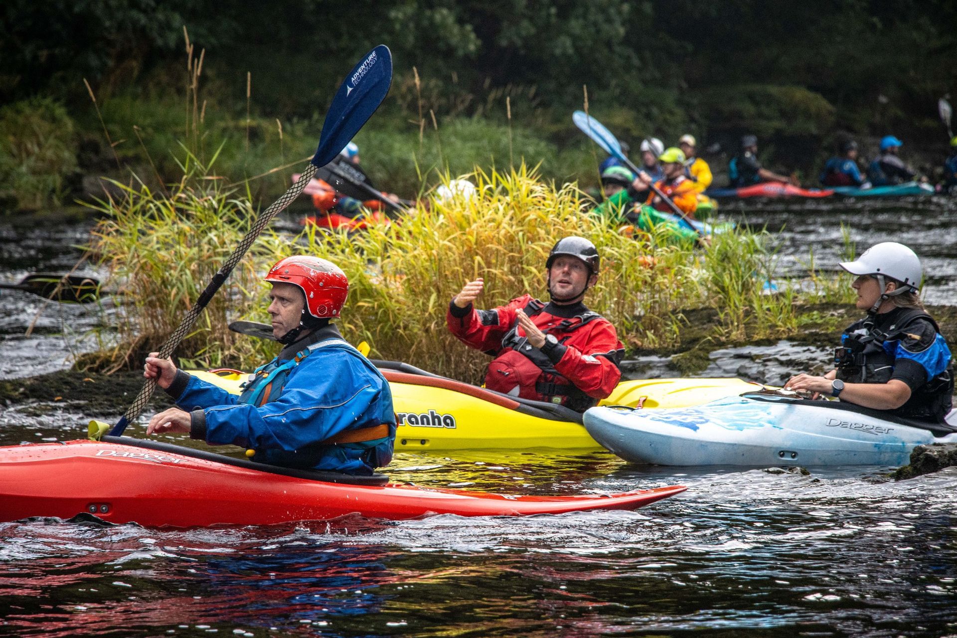Kayakers navigating a river with rapids; some wear helmets, paddles in hand.
