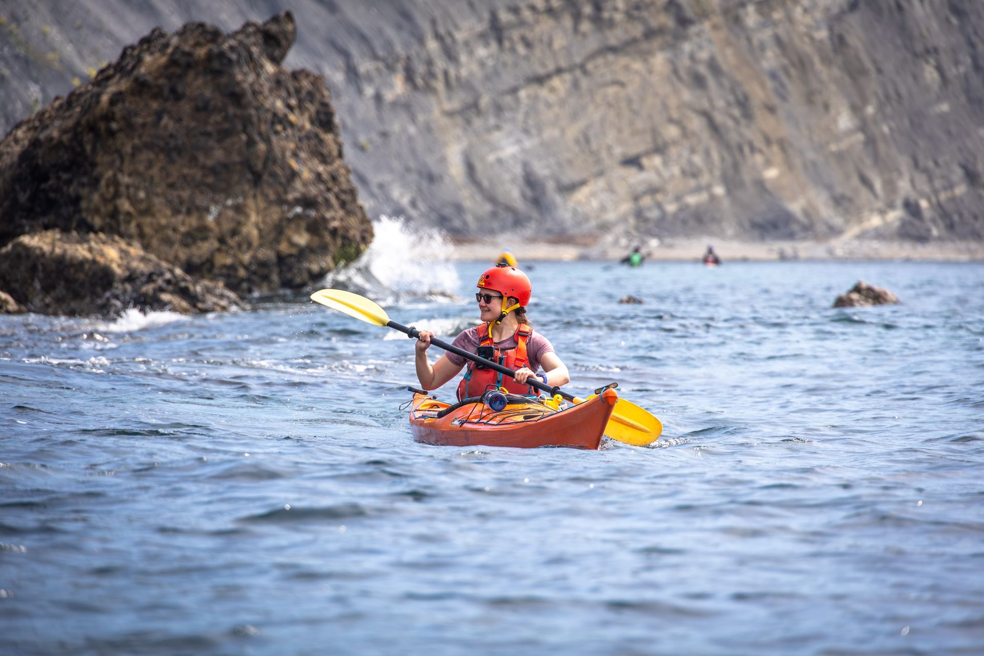 Kayaker in red helmet paddling a yellow kayak on calm water near rocky cliffs