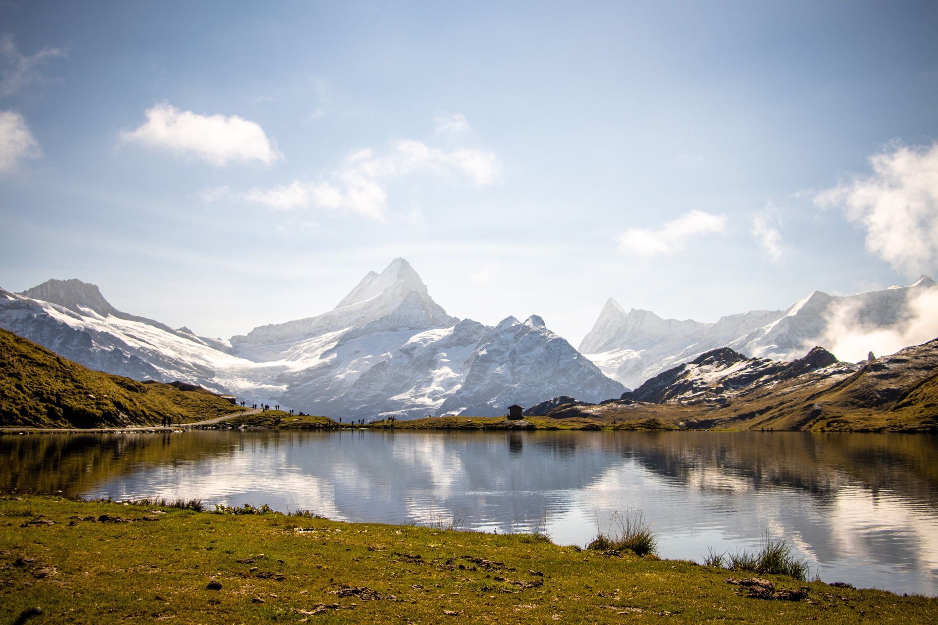 Snow-capped mountains reflected in calm lake under a partly cloudy blue sky. Green grass in the foreground.