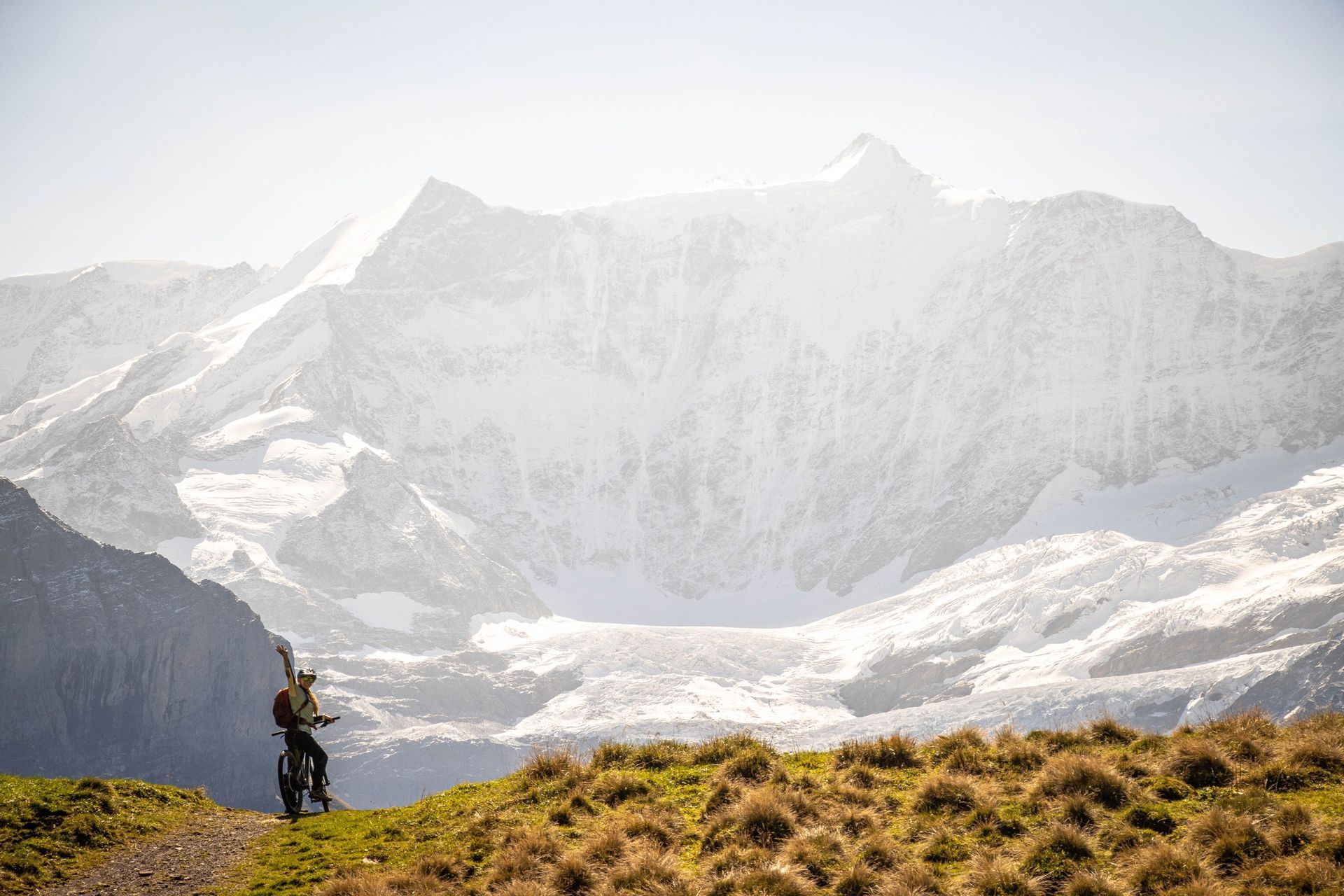 Mountain biker carrying bike on a trail, snowy mountains in background, sunny.