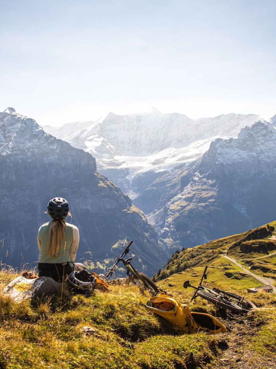 Woman in helmet gazes at snow-capped mountains; mountain bikes rest nearby on a sunny hillside.