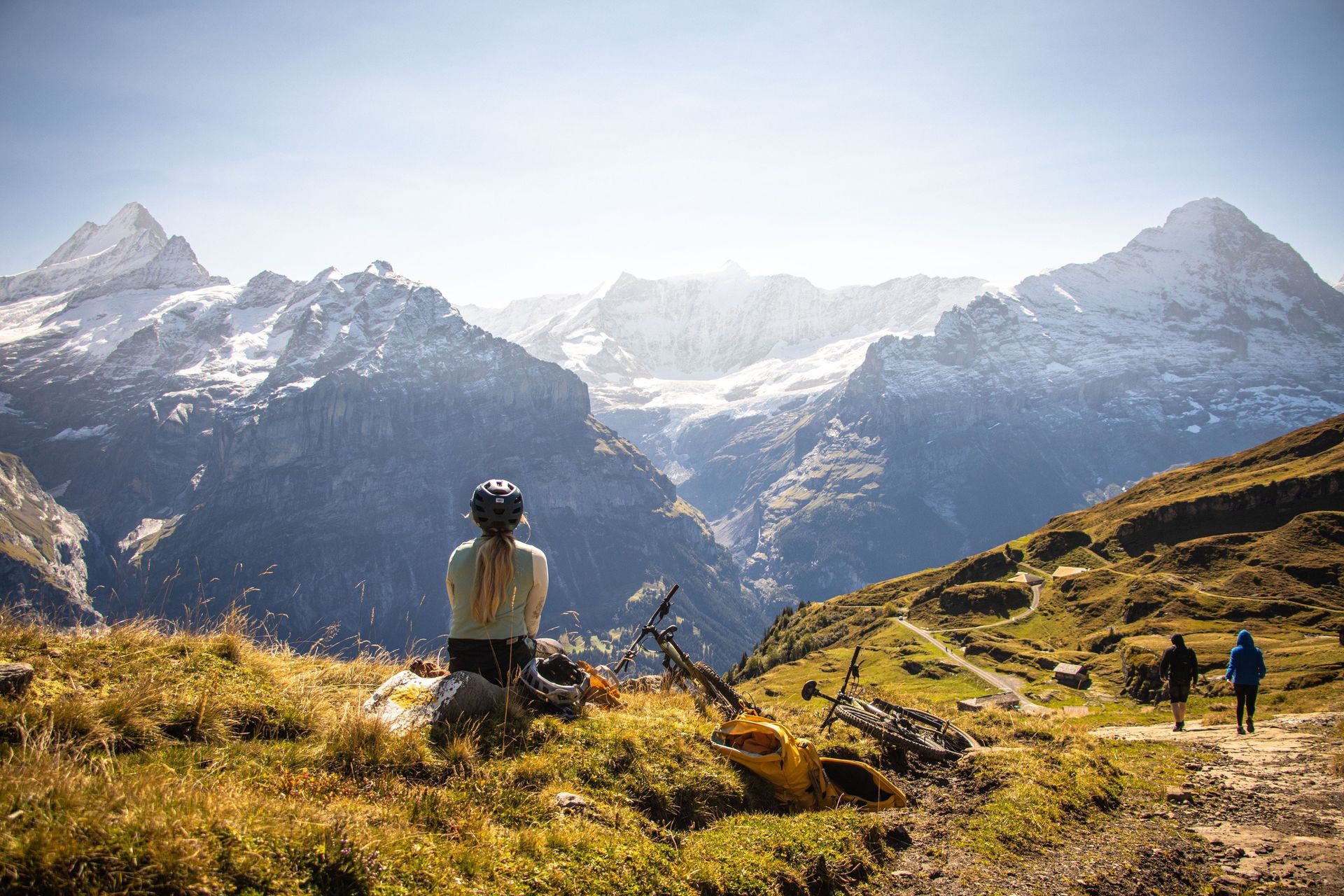 Person seated, looking at snowy mountains, bicycle nearby. Two people walk on a path in the Swiss Alps.