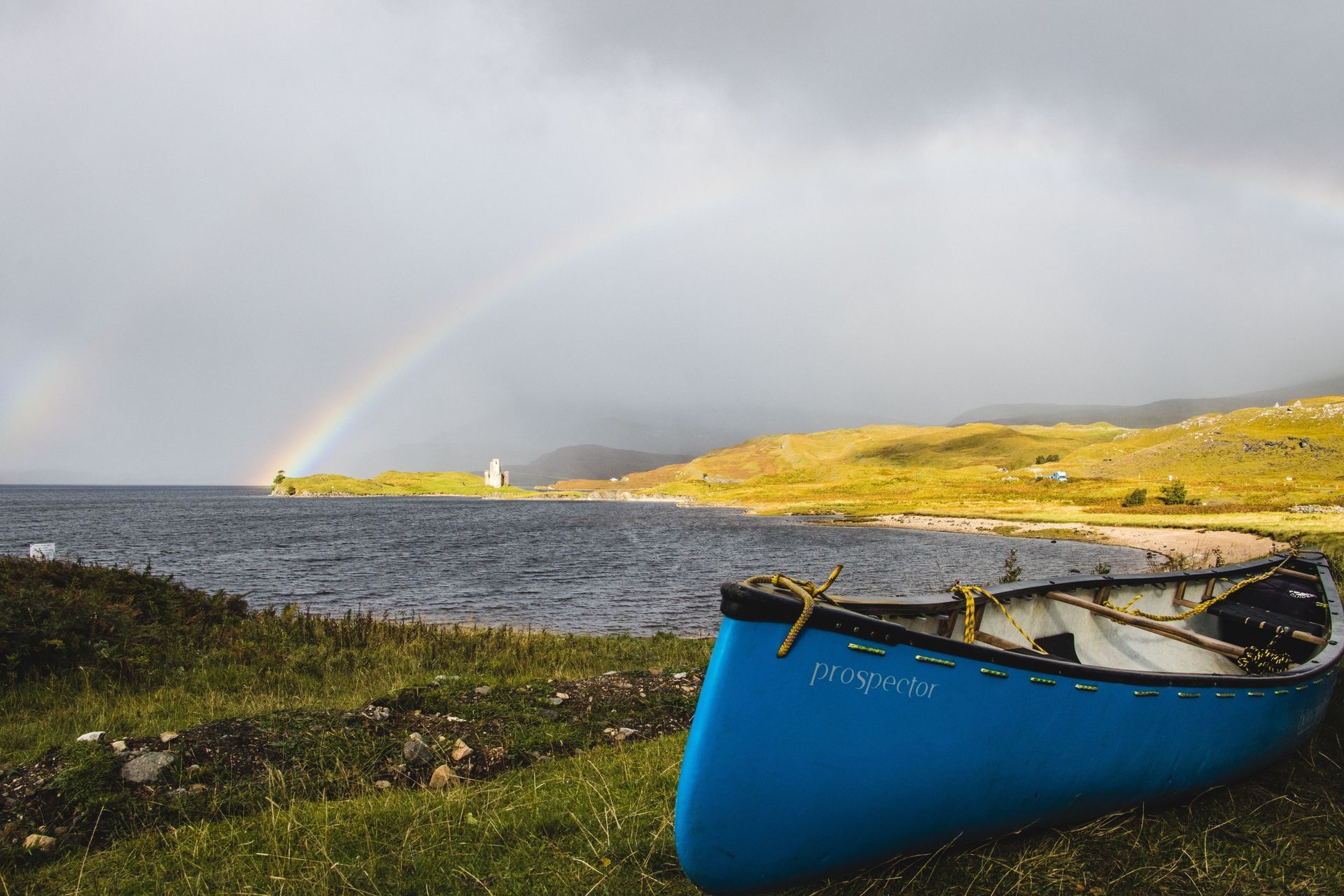 Blue canoe on shore with a rainbow over a lake and distant landscape.
