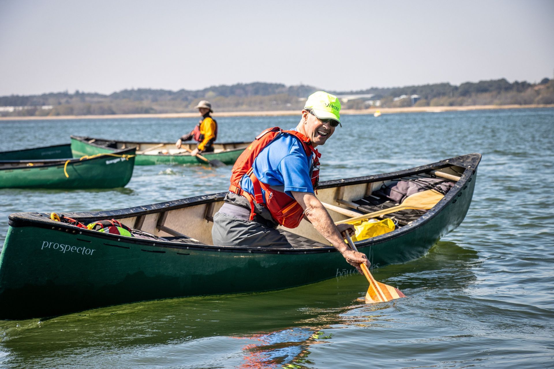 Man paddling a green canoe on water, wearing a life vest and a hat. Another canoe is in the background.