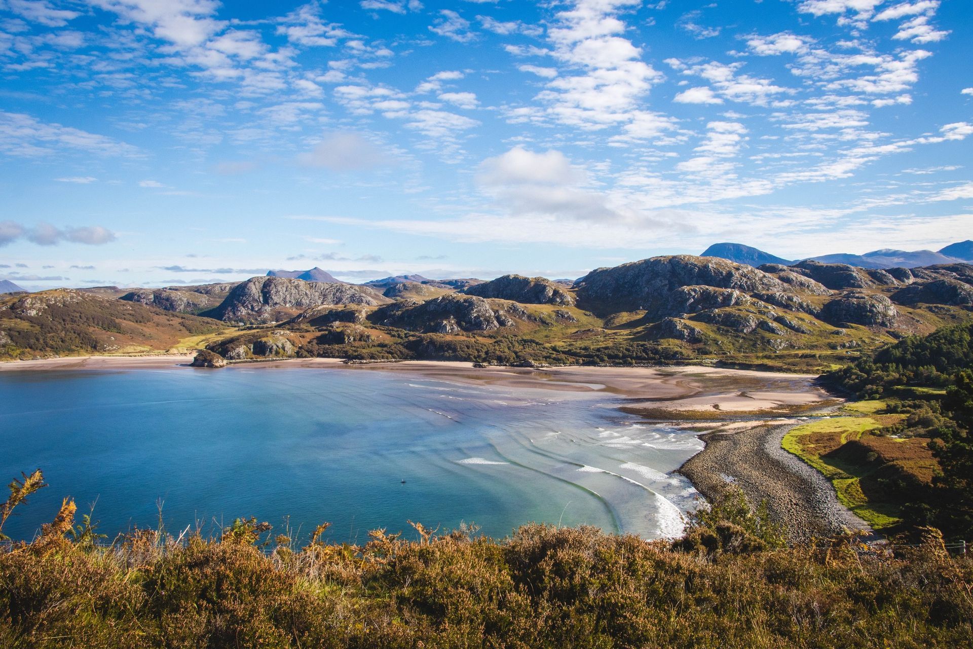 Bay with blue water, sandy beach, and rocky hills under a partly cloudy sky.