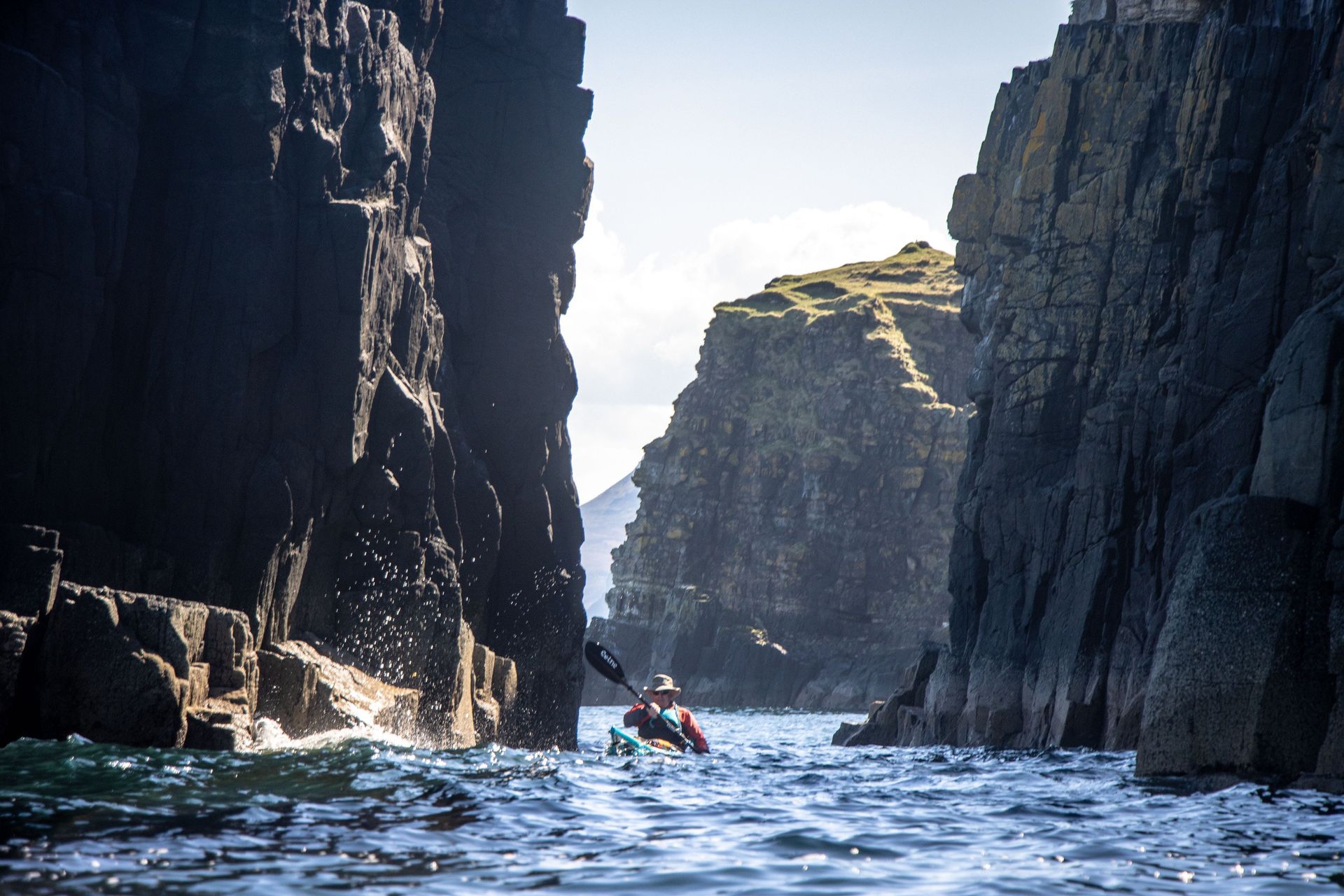 Kayaker paddling through a narrow sea canyon with high, dark rock walls, clear water, and sunny sky.