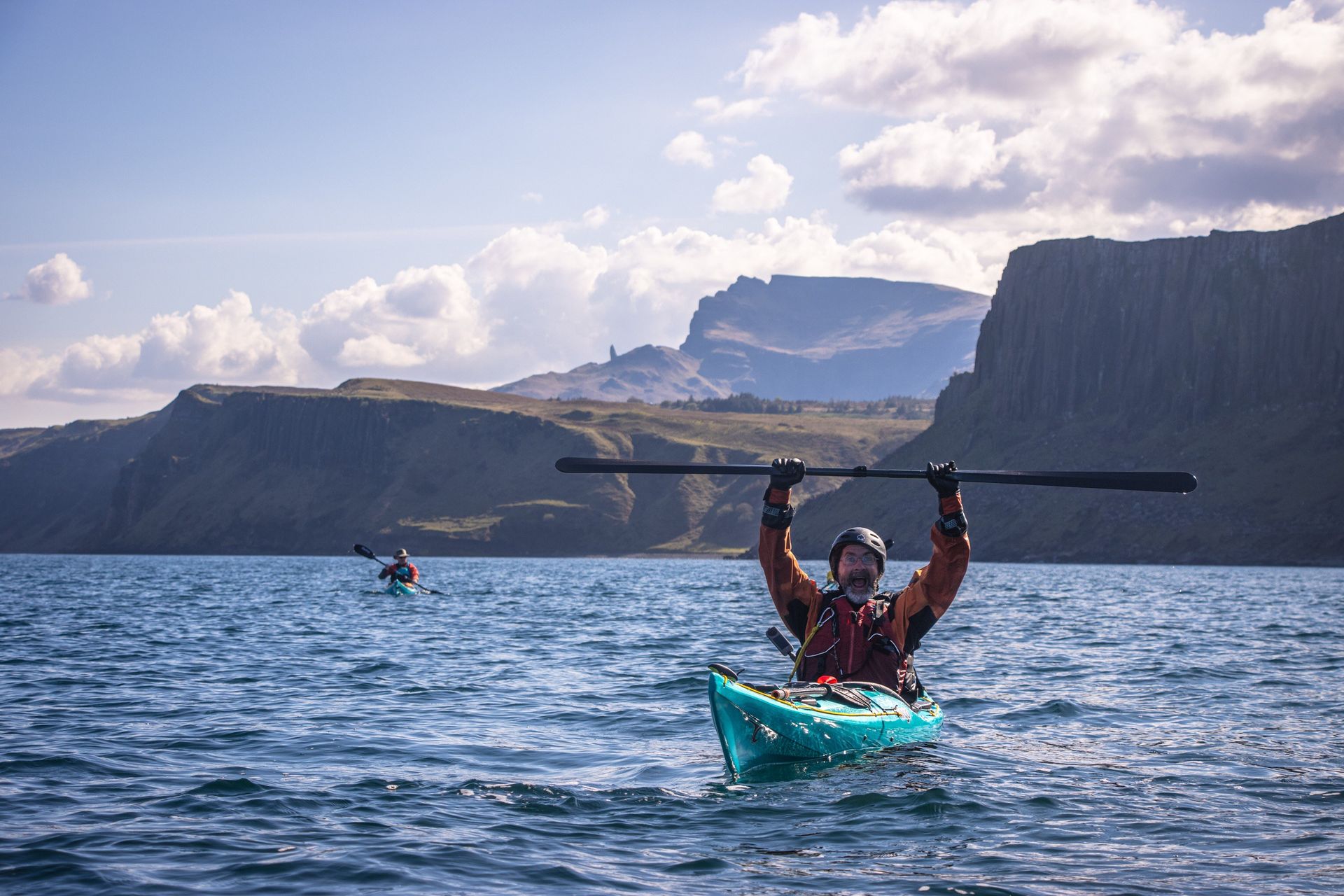 Man in kayak raises paddle, blue water, cliffs in background, sunny day.