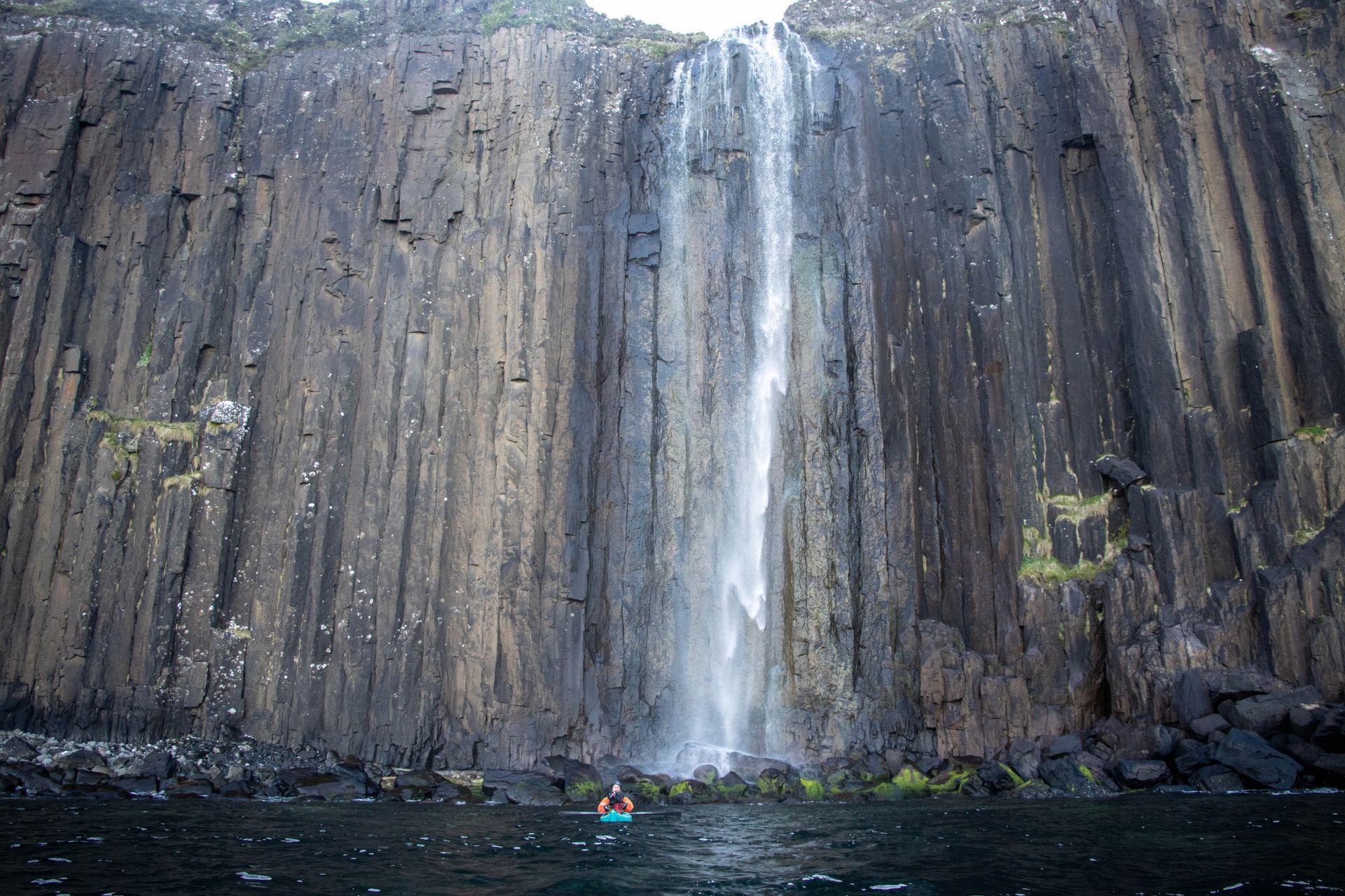 Waterfall cascading down a dark, columnar cliff face; a person kayaks in the water below.