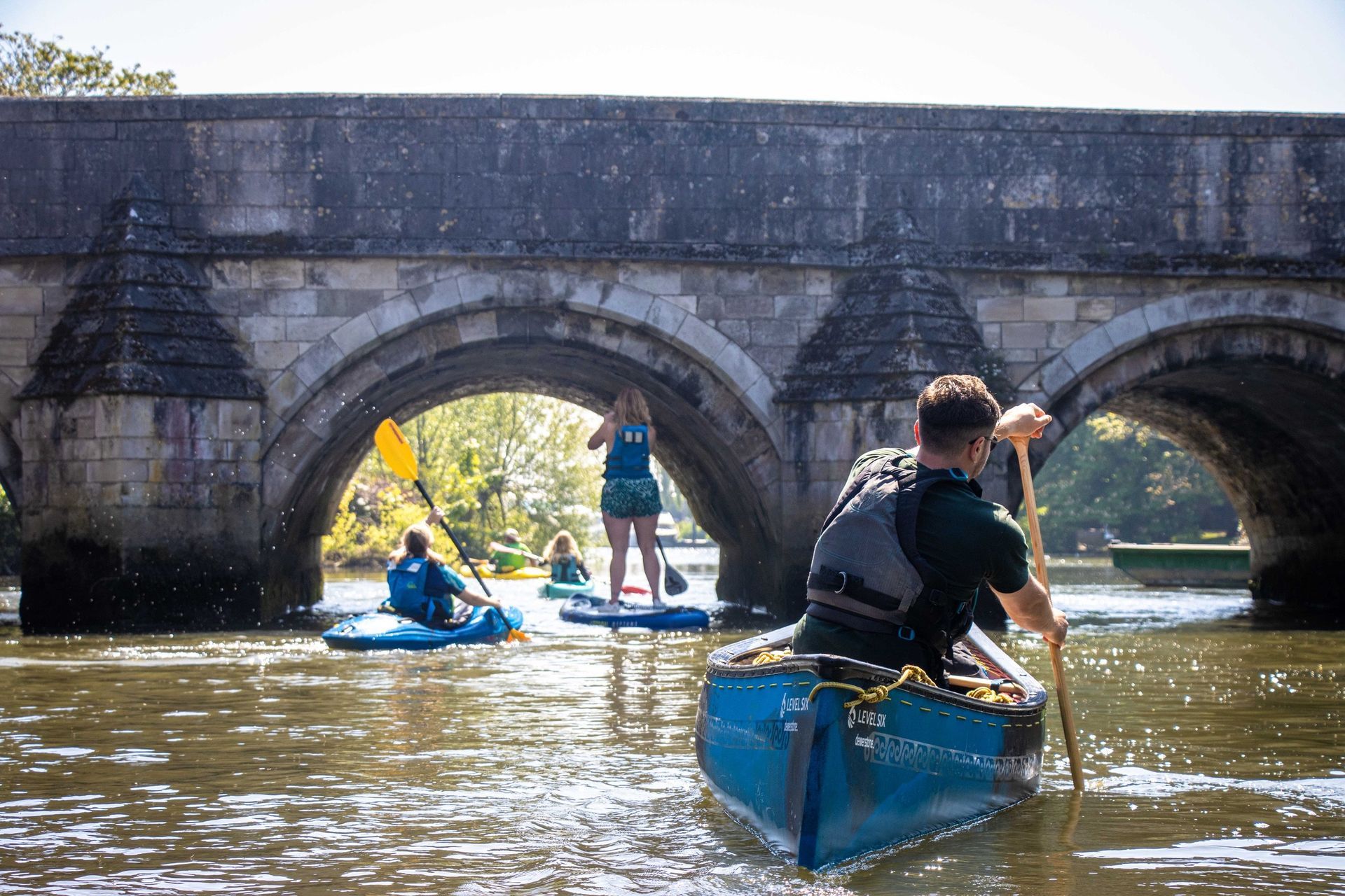 People kayaking and paddleboarding under a stone bridge on a sunny day.