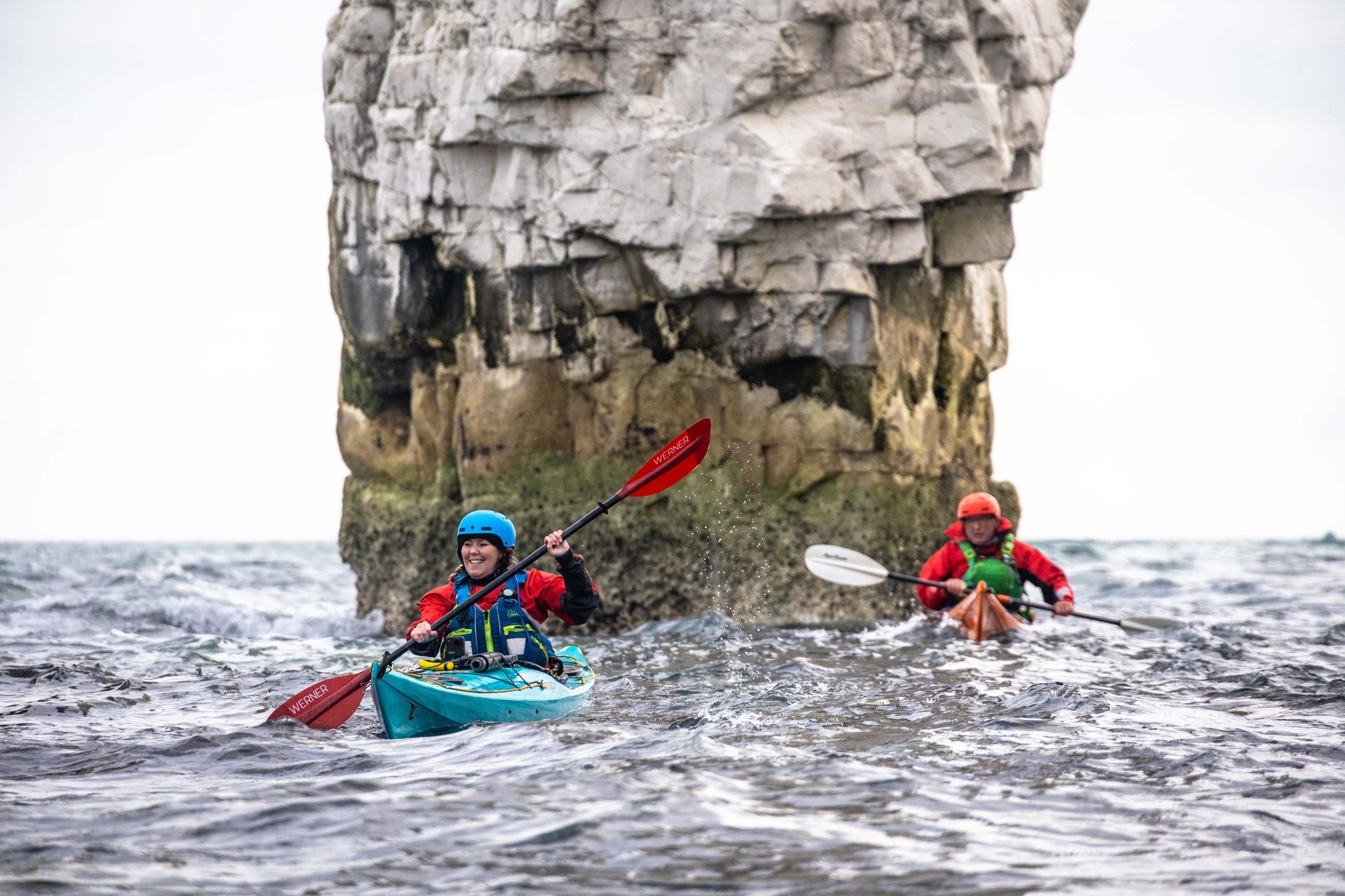 Two people kayaking near a large rock formation in choppy water, one raising their paddle.