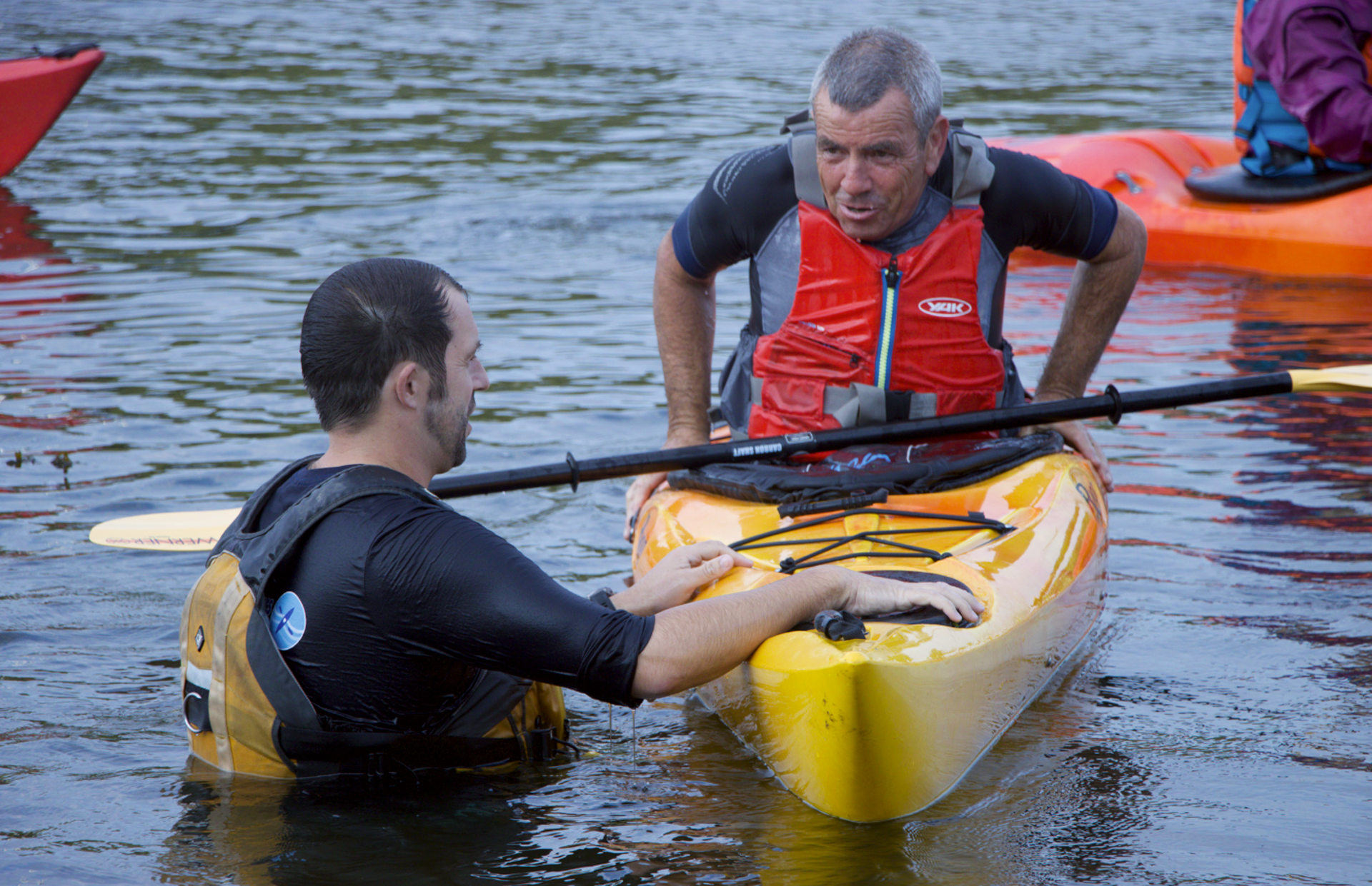 Two men in a lake. One in a kayak, other helping from water. Both wearing life vests.