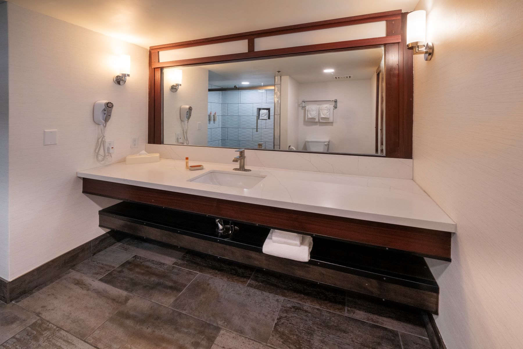 Bathroom with a long white countertop, large mirror, and open shelf with towels.