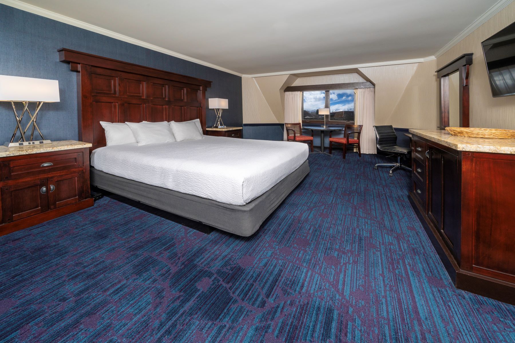 Hotel bedroom with a large bed, blue carpet, dark wood furniture, and a window.