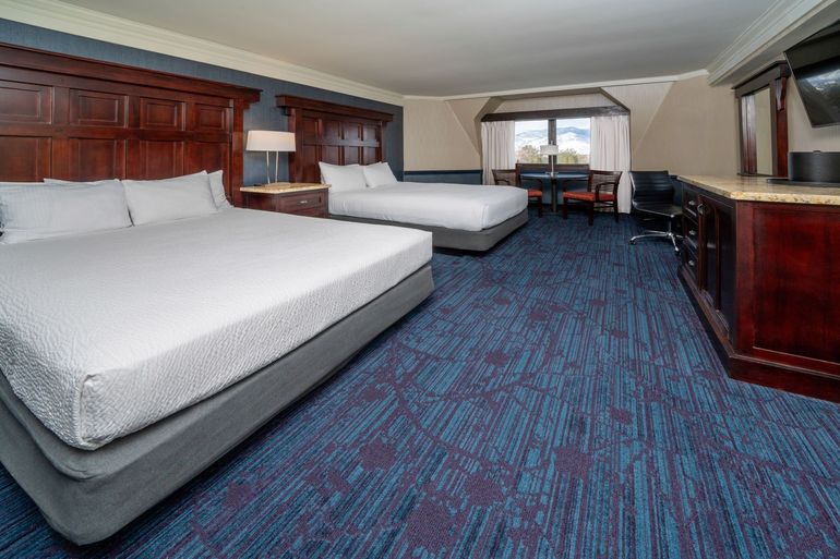 Hotel room with two queen beds, a table, and a TV; dark wood furniture, blue carpet.