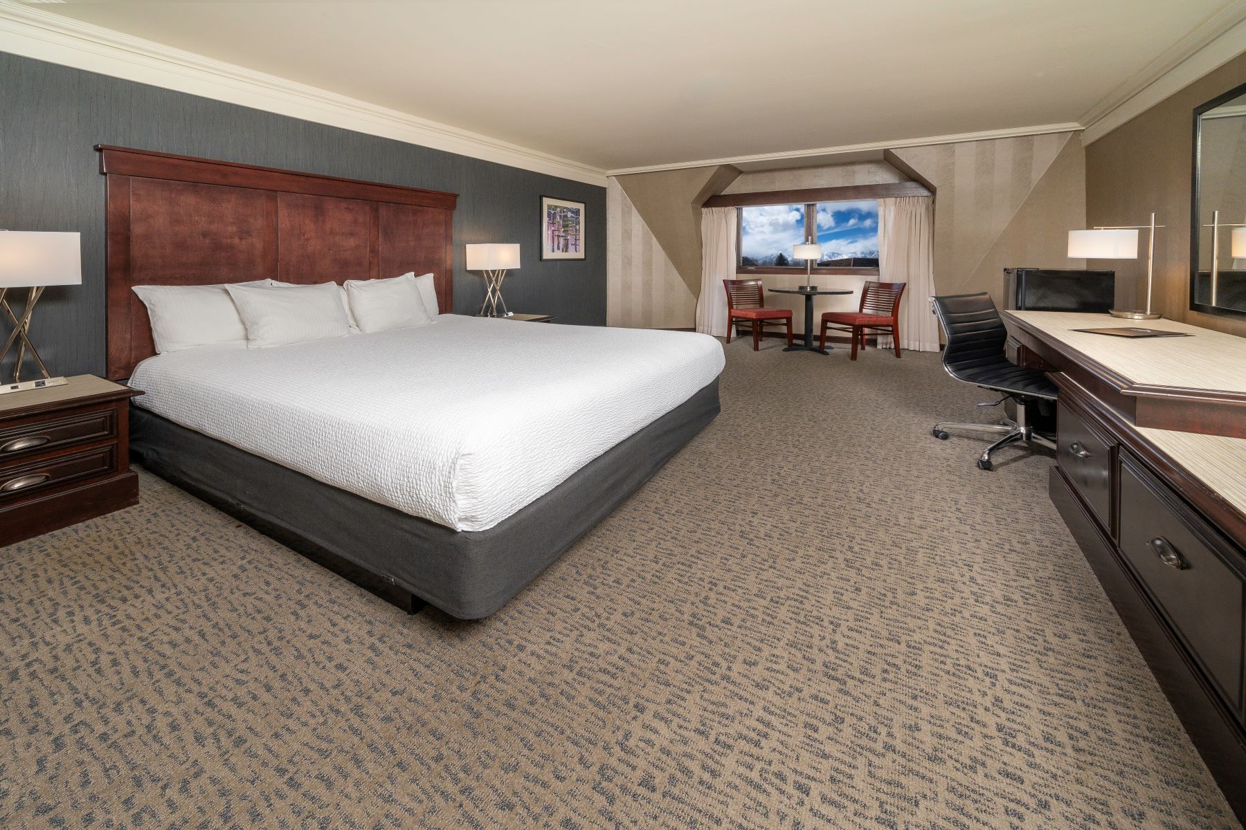 Bedroom with king bed, desk, and small seating area by window. Dark brown furniture and patterned carpet.