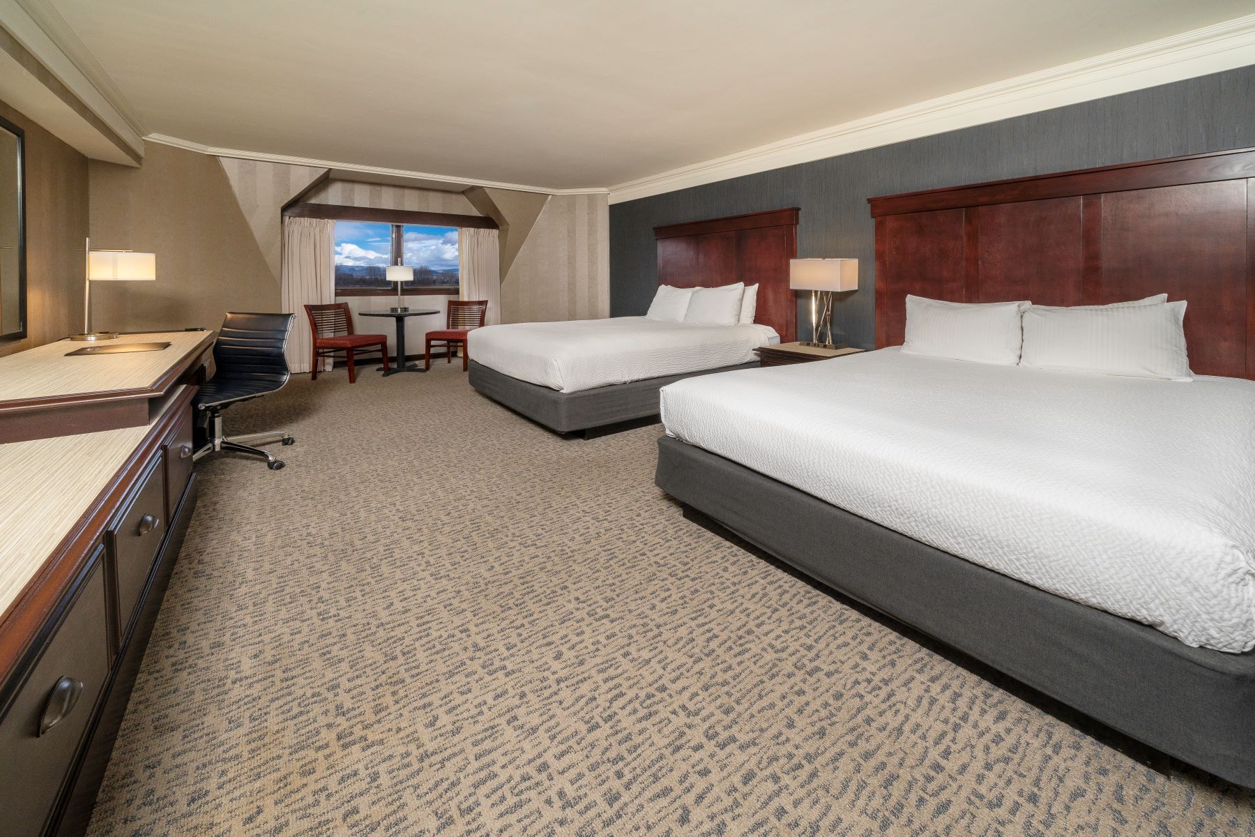 Hotel room with two queen beds, desk, and small table with chairs near a window.
