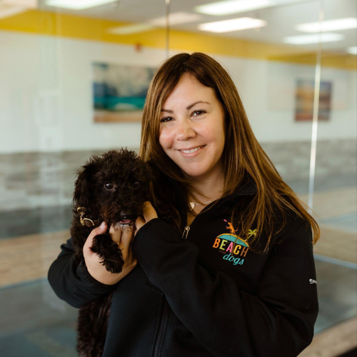 Woman in black sweatshirt holding a small black puppy indoors, smiling at the camera.