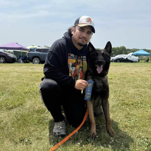 Person kneeling with a German Shepherd on a grassy field, holding a blue ribbon at an outdoor event