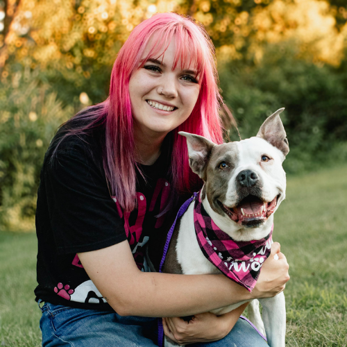 Smiling person with pink hair hugging a gray-and-white dog wearing a pink bandana outdoors