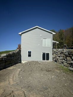 Gray, two-story house with white trim, set between two stone walls and dirt ground, under a blue sky.