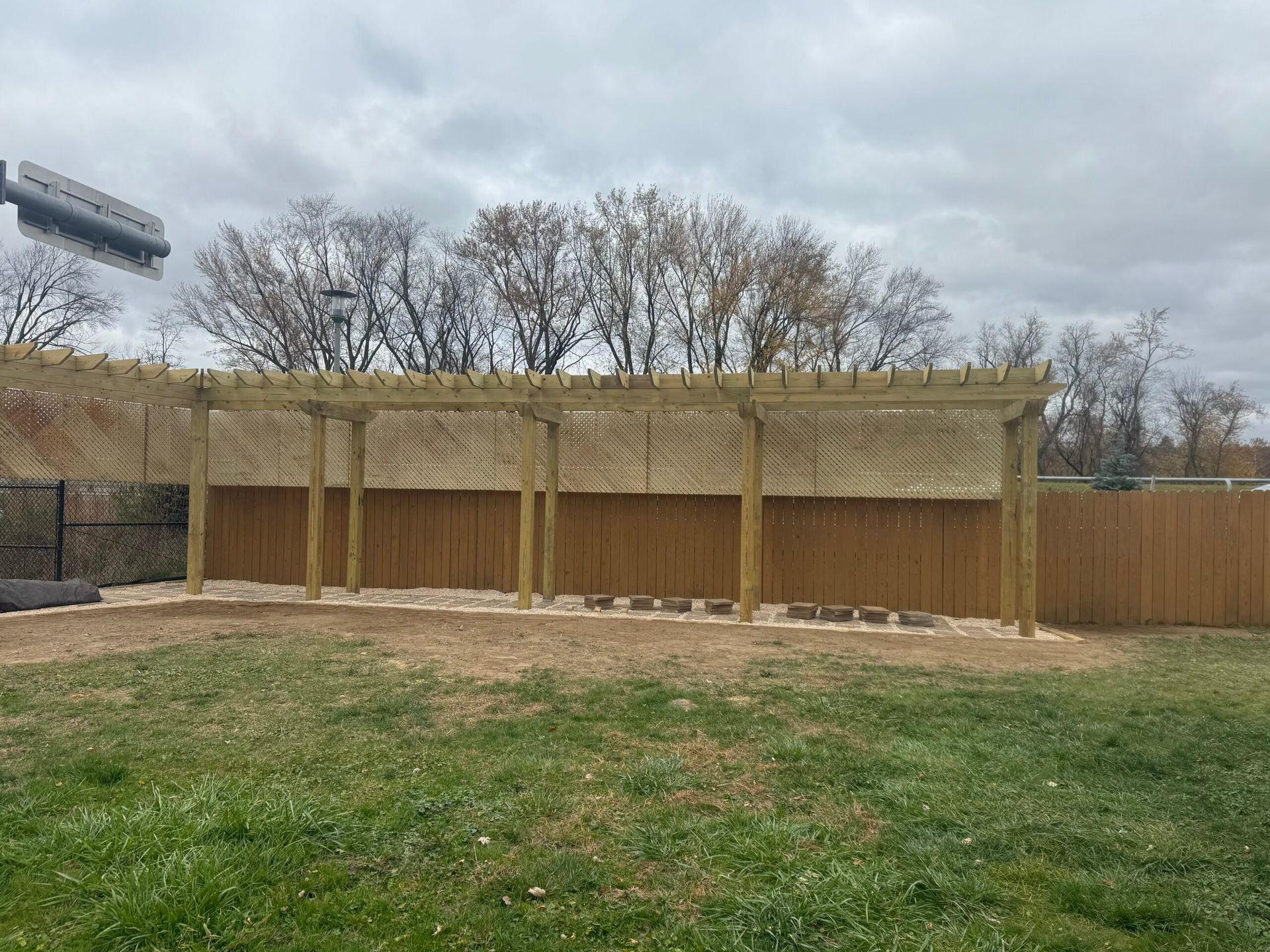 A wooden pergola with posts stands in a yard against a wooden fence on an overcast day.