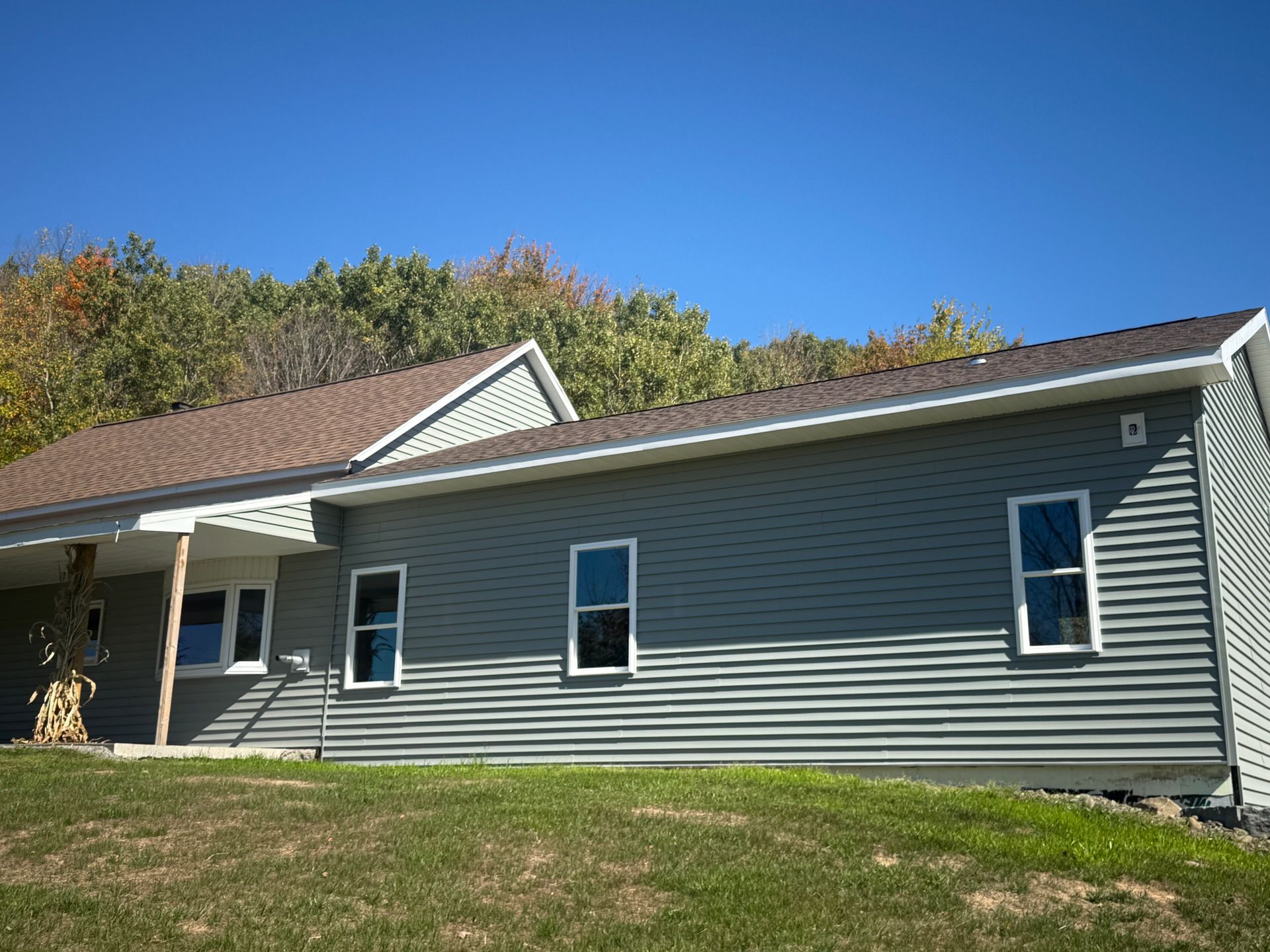 Gray house with three windows and brown roof against a blue sky, trees in the background.