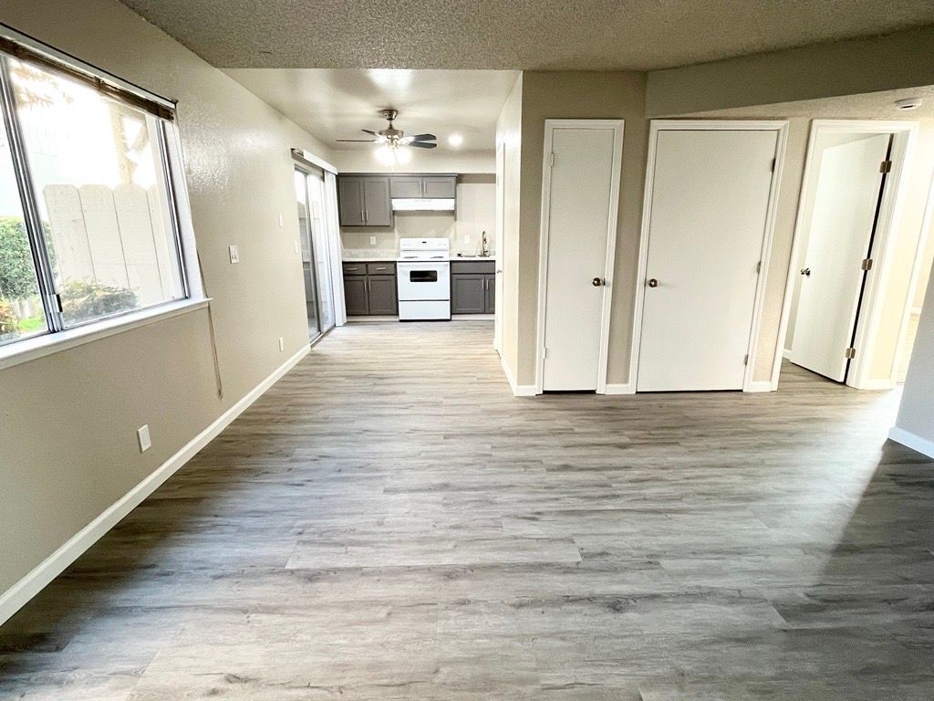 Living room with wood-look floor, kitchen in background; natural light, neutral walls, white appliances, two white doors.