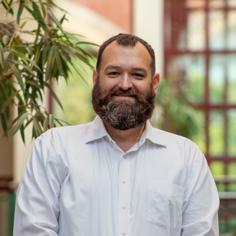 Smiling bearded man in a white shirt standing indoors with plants and windows behind him
