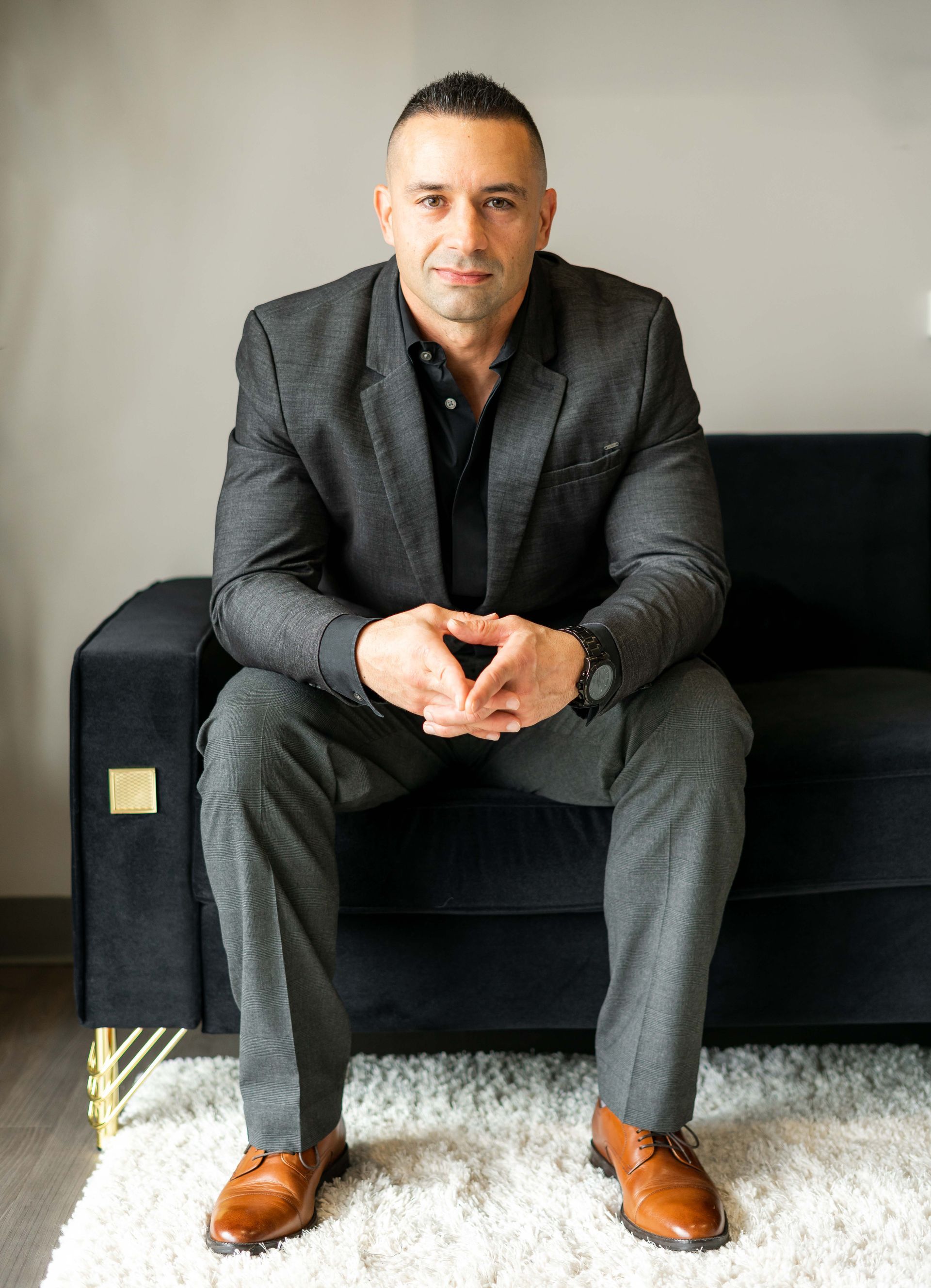 Man in a dark suit seated on a black sofa, leaning forward with hands clasped in a bright room