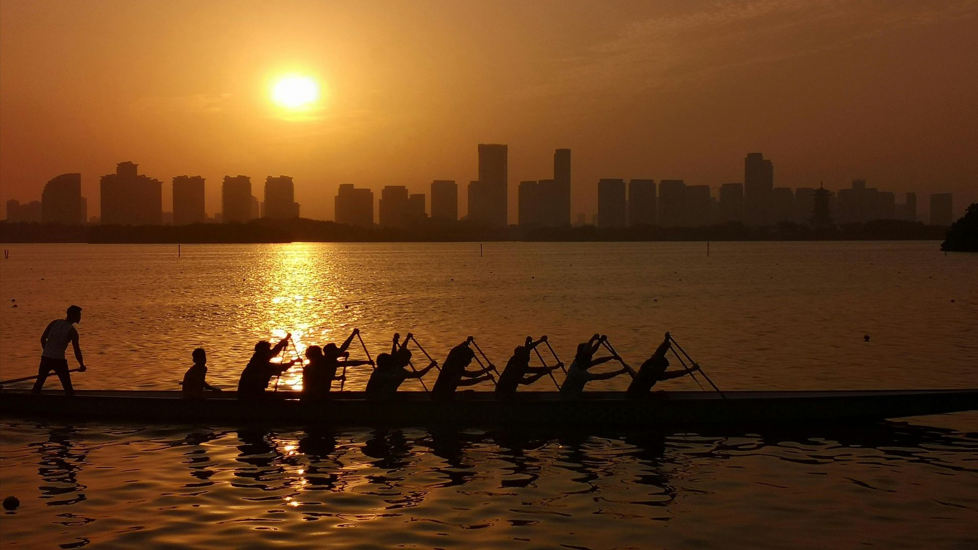 Rowers silhouetted on a boat at sunset, with a glowing city skyline across the water.