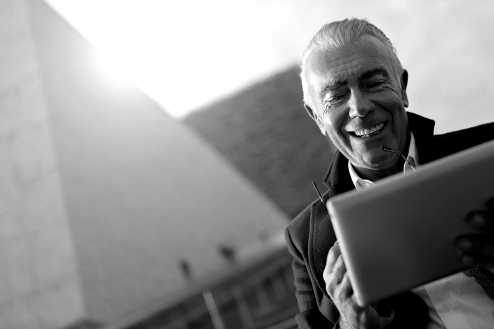 Smiling person holding a tablet outdoors in black and white, with a building in the background
