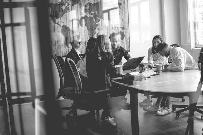 People gathered around a round table in a bright office meeting room, discussing over laptops and papers.
