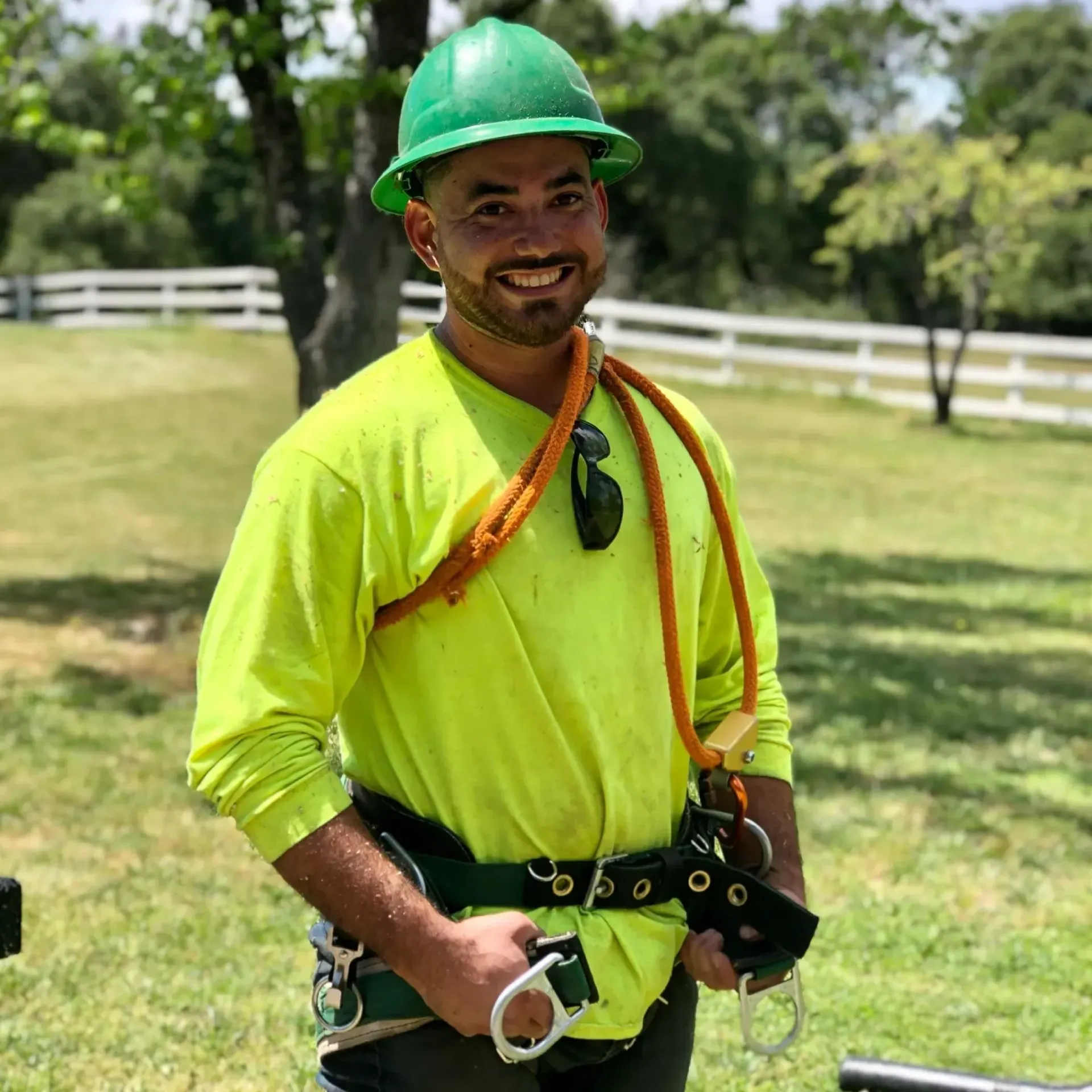 Man in lime green work shirt and hard hat, wearing a safety harness, smiling outside.